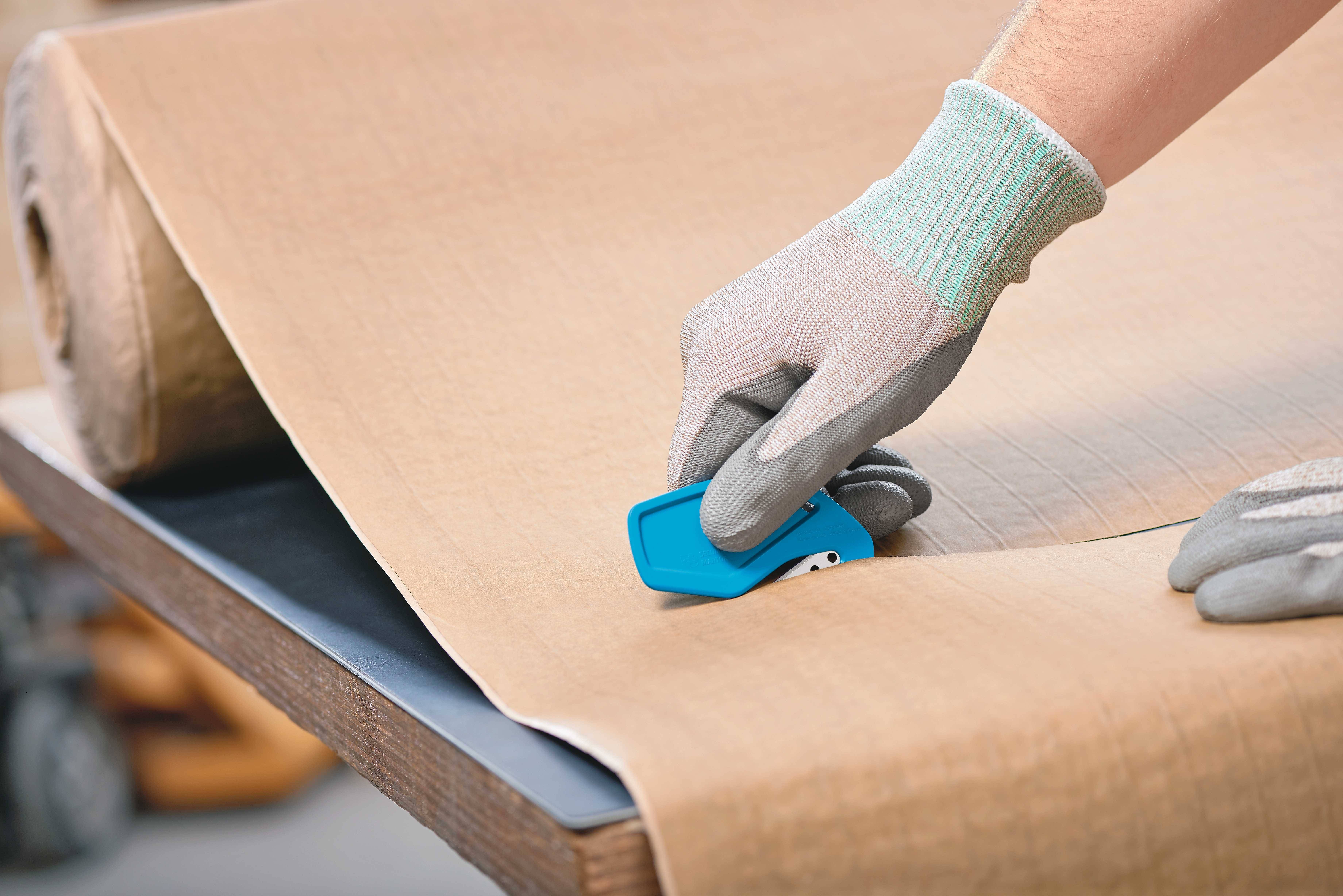 Une personne portant des gants coupe un grand rouleau de papier brun sur une table à l'aide d'un outil de coupe bleu.