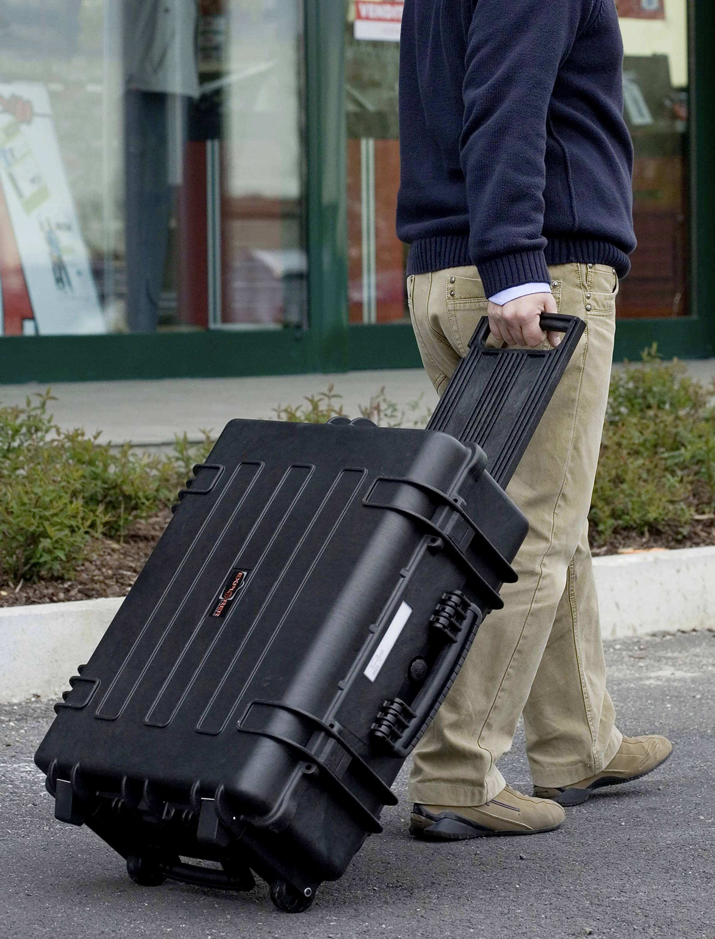 Une personne tire une grande valise rigide noire à roulettes par sa poignée sur un parking devant un bâtiment.