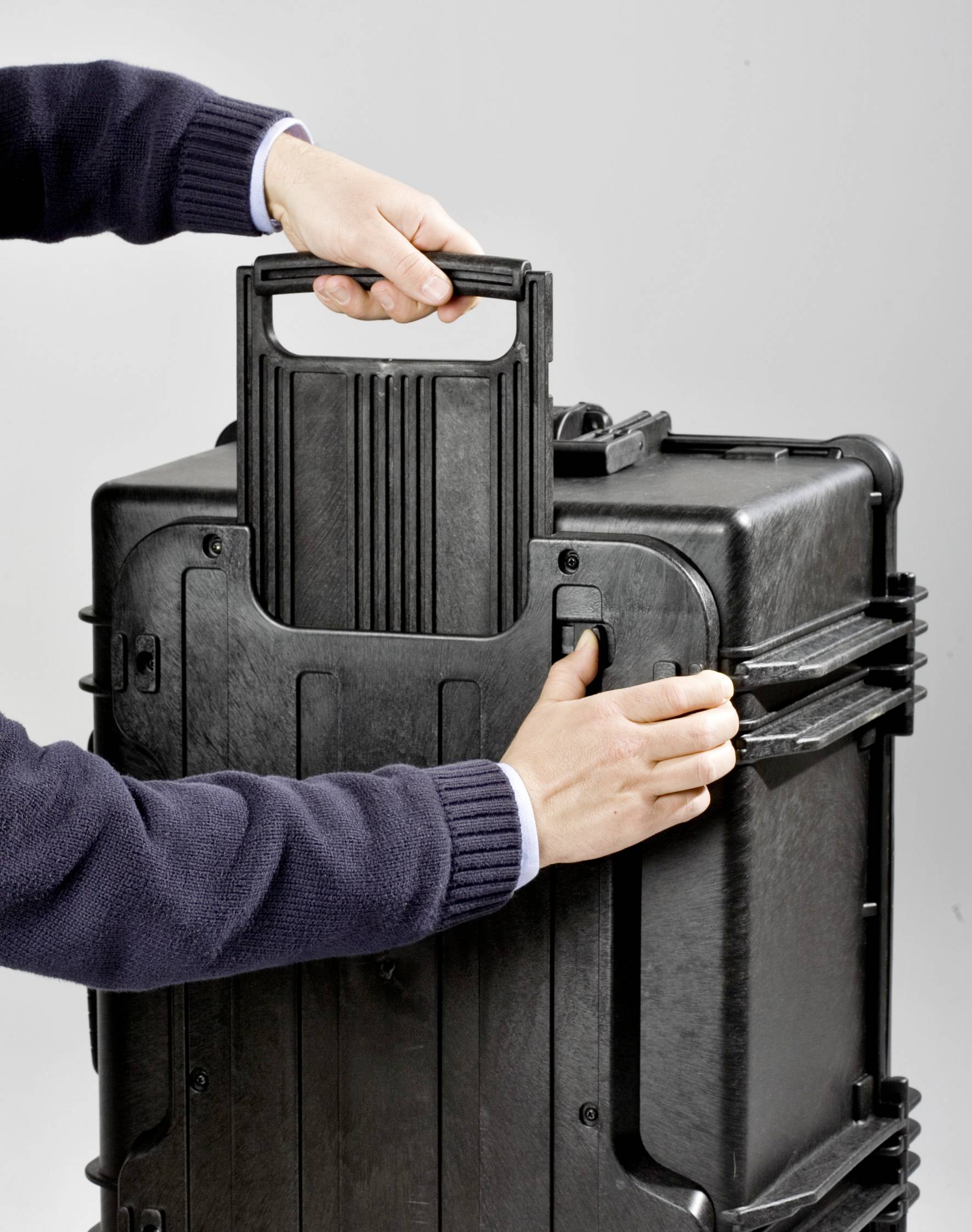 Une personne soulève une valise de transport robuste et noire par sa poignée. La valise possède des coins renforcés et est conçue pour supporter des charges lourdes.