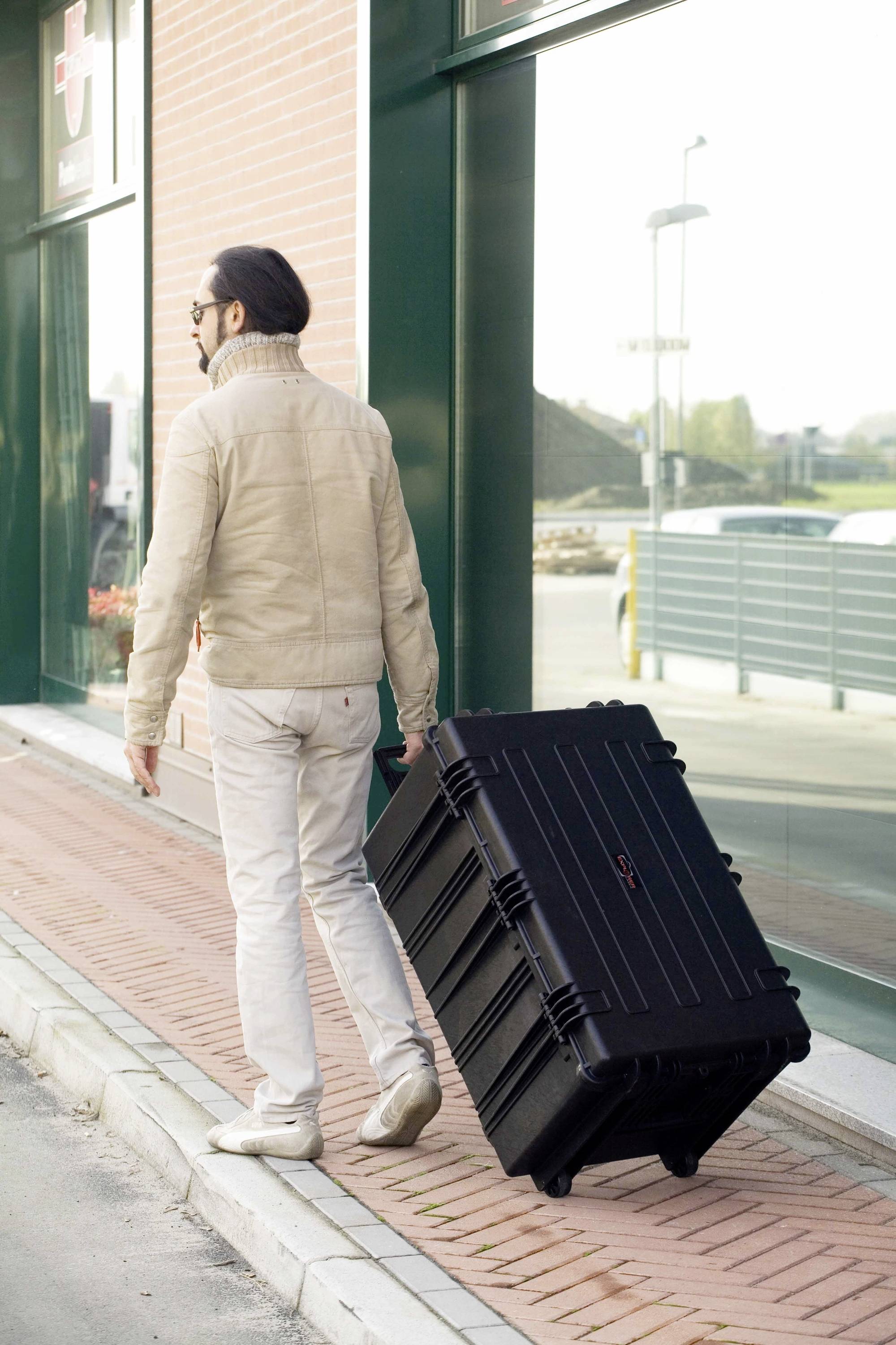 Une personne vêtue de vêtements clairs tire une grande valise noire sur le trottoir le long d'une façade vitrée.