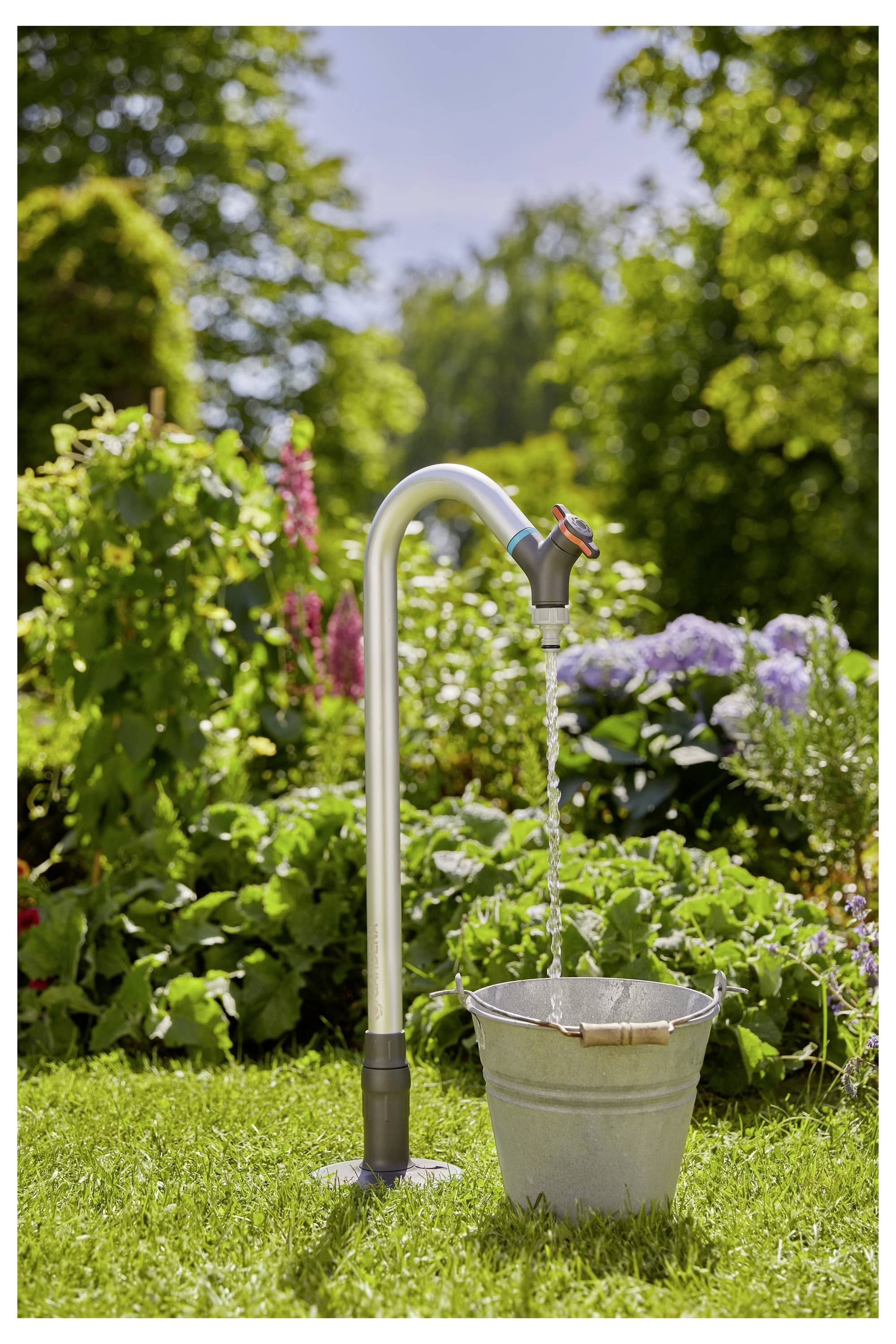Un robinet en métal avec de l'eau qui coule dans un seau sur une zone herbeuse, entouré de plantes de jardin luxuriantes sous un ciel bleu clair.