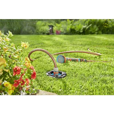 Un tuyau d'arrosage avec un diffuseur est posé sur une pelouse verte. À côté se trouvent des parterres de fleurs. La scène illustre l'entretien d'un jardin.