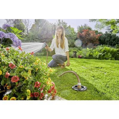 Une femme souriante arrose des fleurs colorées avec un tuyau d'arrosage dans un jardin ensoleillé. Des arbres sont visibles en arrière-plan.