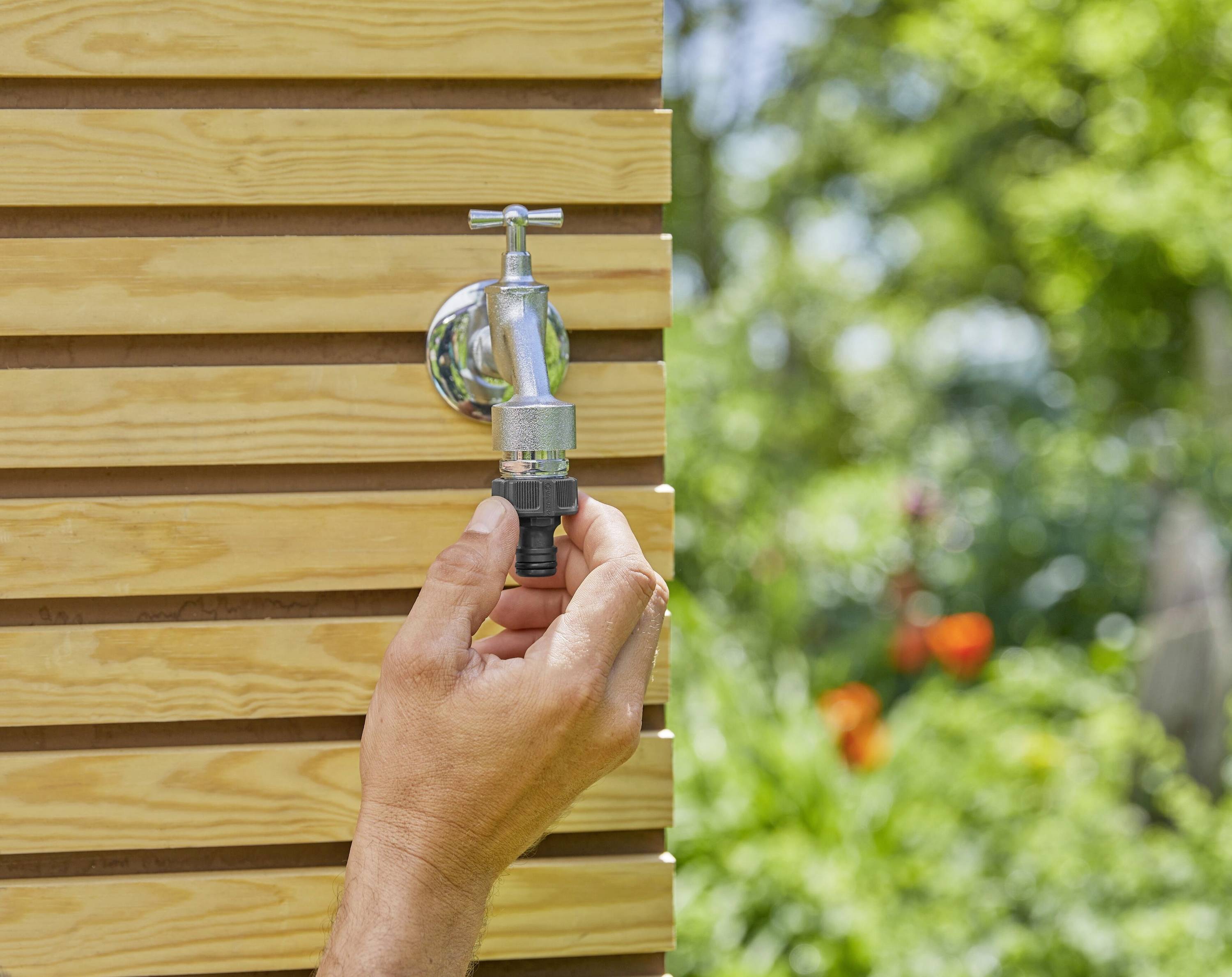 Une main tourne un robinet sur une structure en bois à l'extérieur. Un arrière-plan flou de verdure et de fleurs est visible.