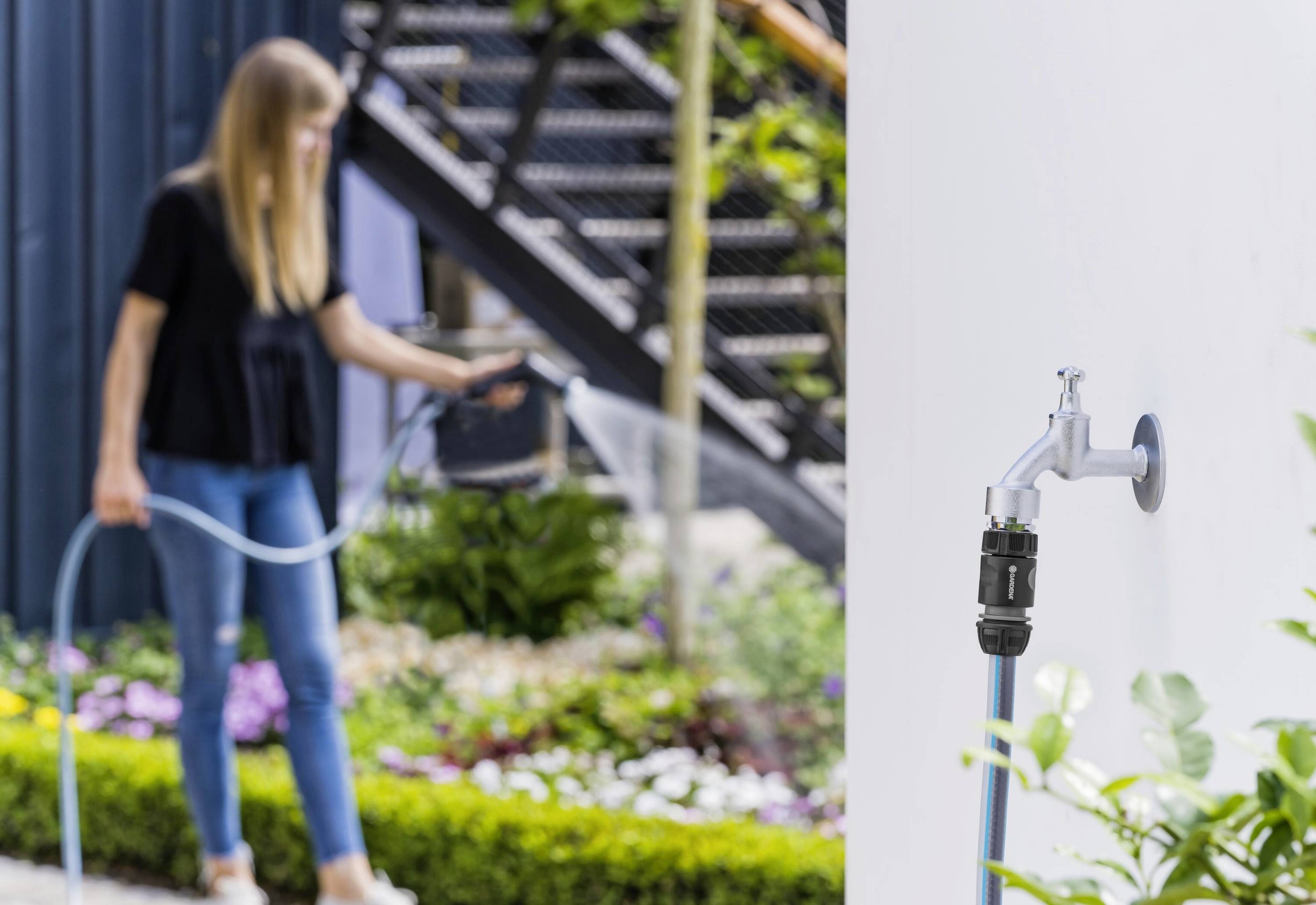 Femme aux longs cheveux blonds arrosant des plantes avec un tuyau d'arrosage. Un robinet est visible au premier plan contre un mur blanc.