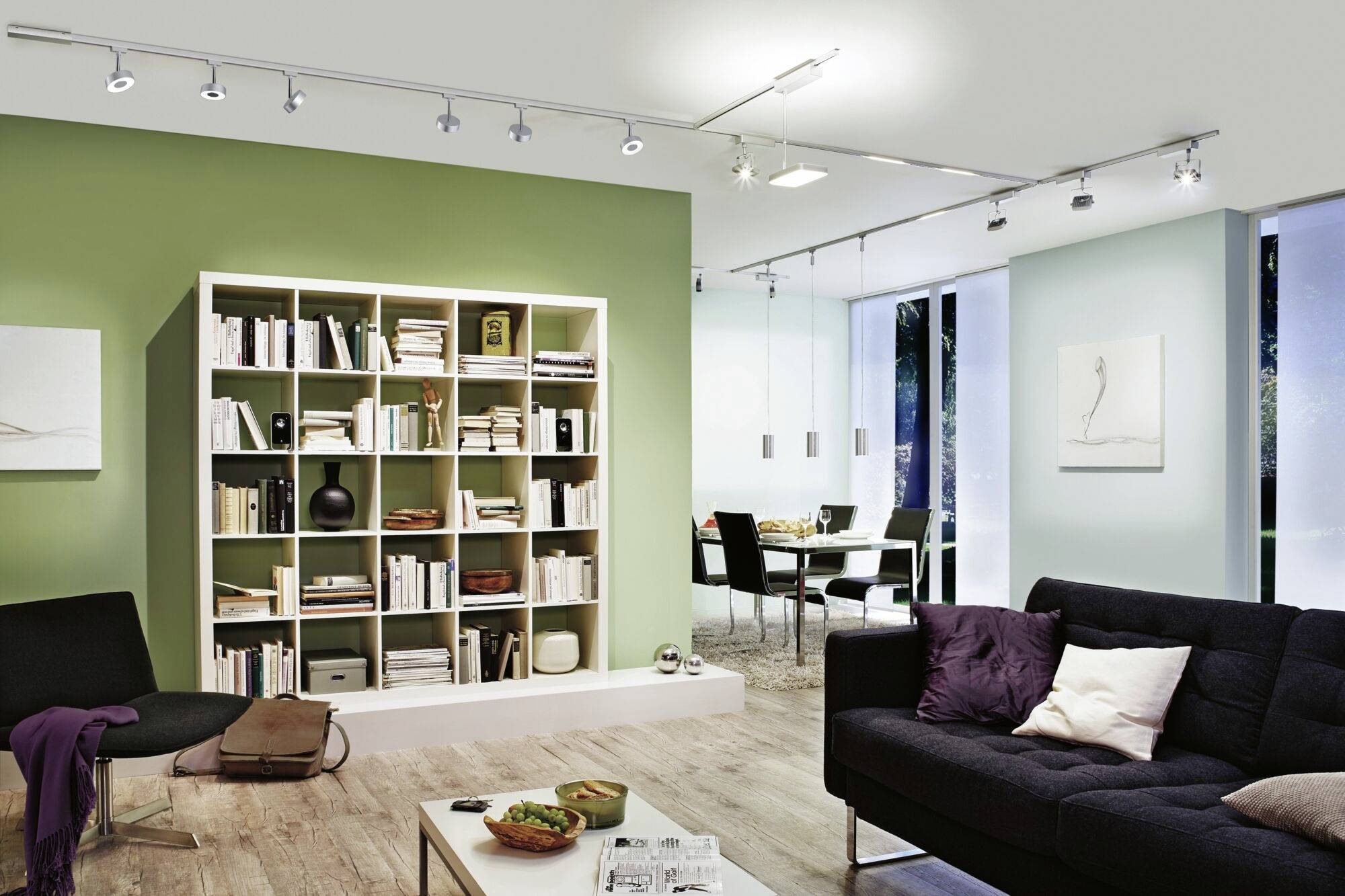 Salon lumineux avec un mur peint en vert, une bibliothèque, un canapé noir et vue sur la salle à manger. Décoration intérieure moderne.