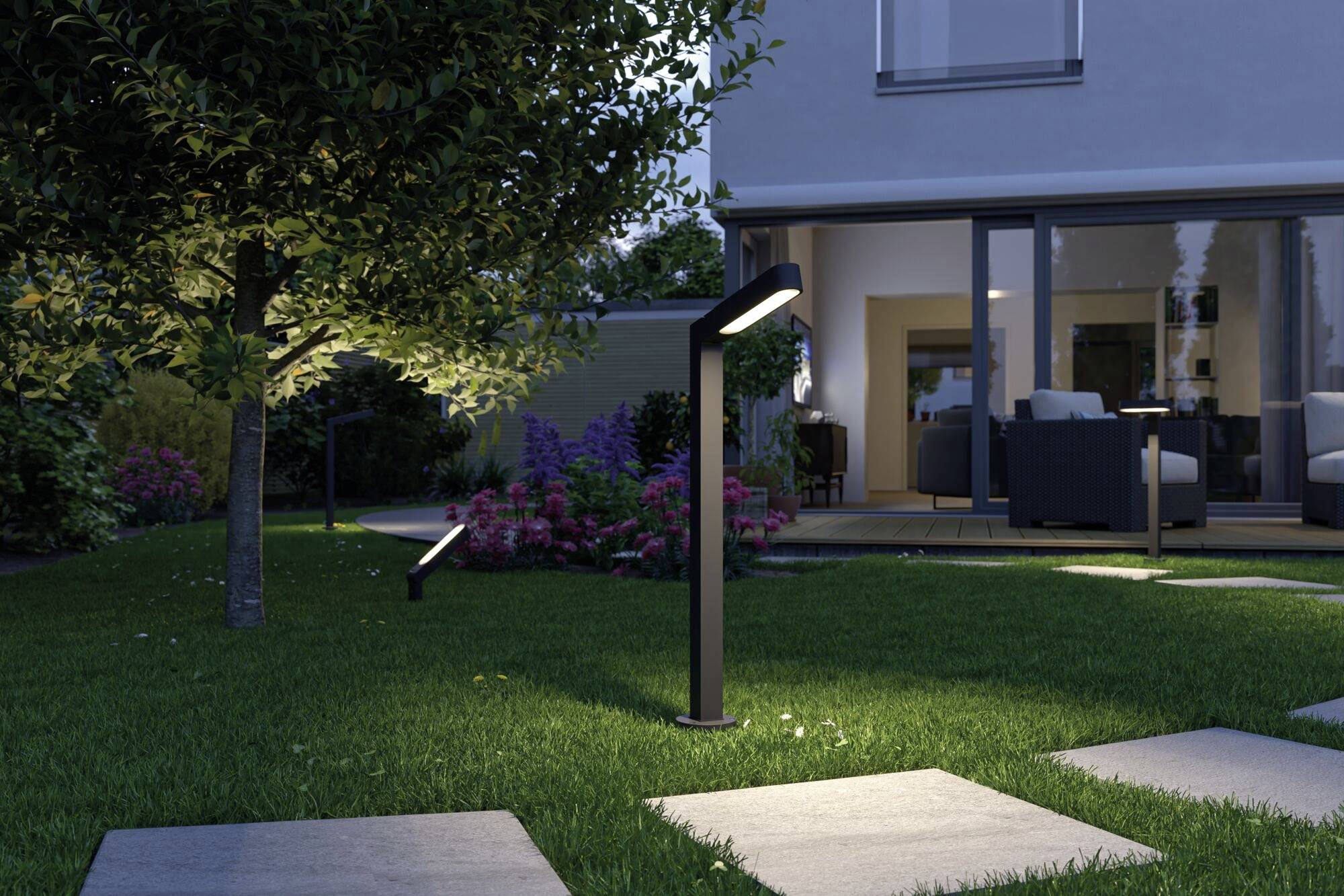 Jardin moderne au crépuscule avec des chemins éclairés, pelouse, arbres et vue sur un salon doucement illuminé.