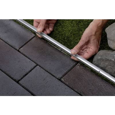 Des mains placent un profilé métallique entre le bord de la pelouse et les pavés. Ceci pourrait faire partie d'un projet de construction ou de rénovation.