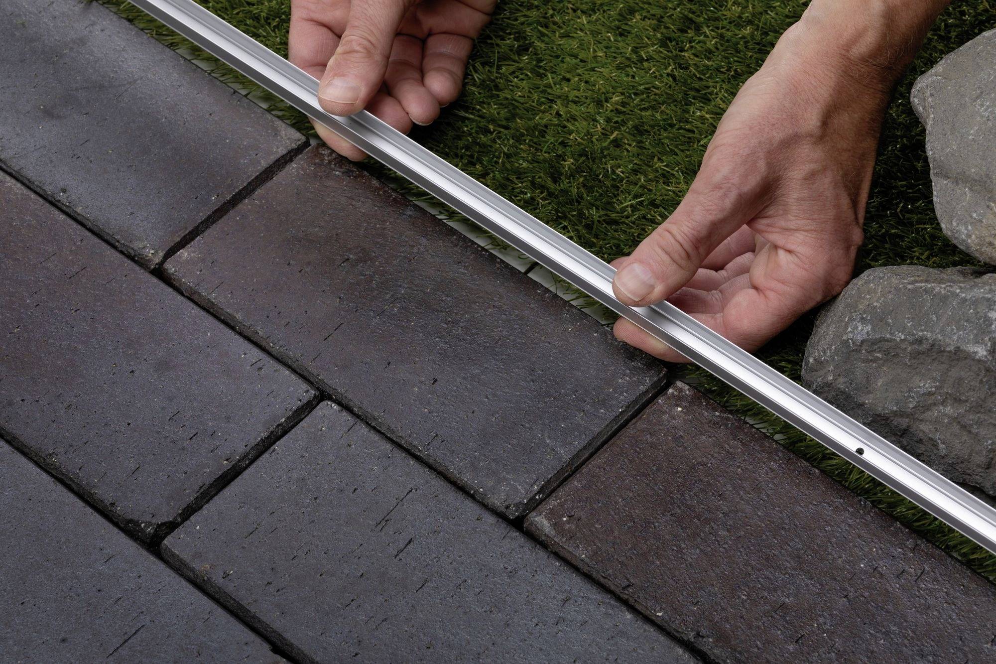 Une personne aligne une barre métallique sur une surface combinant pelouse et pavés pour créer une séparation ou un bord net.