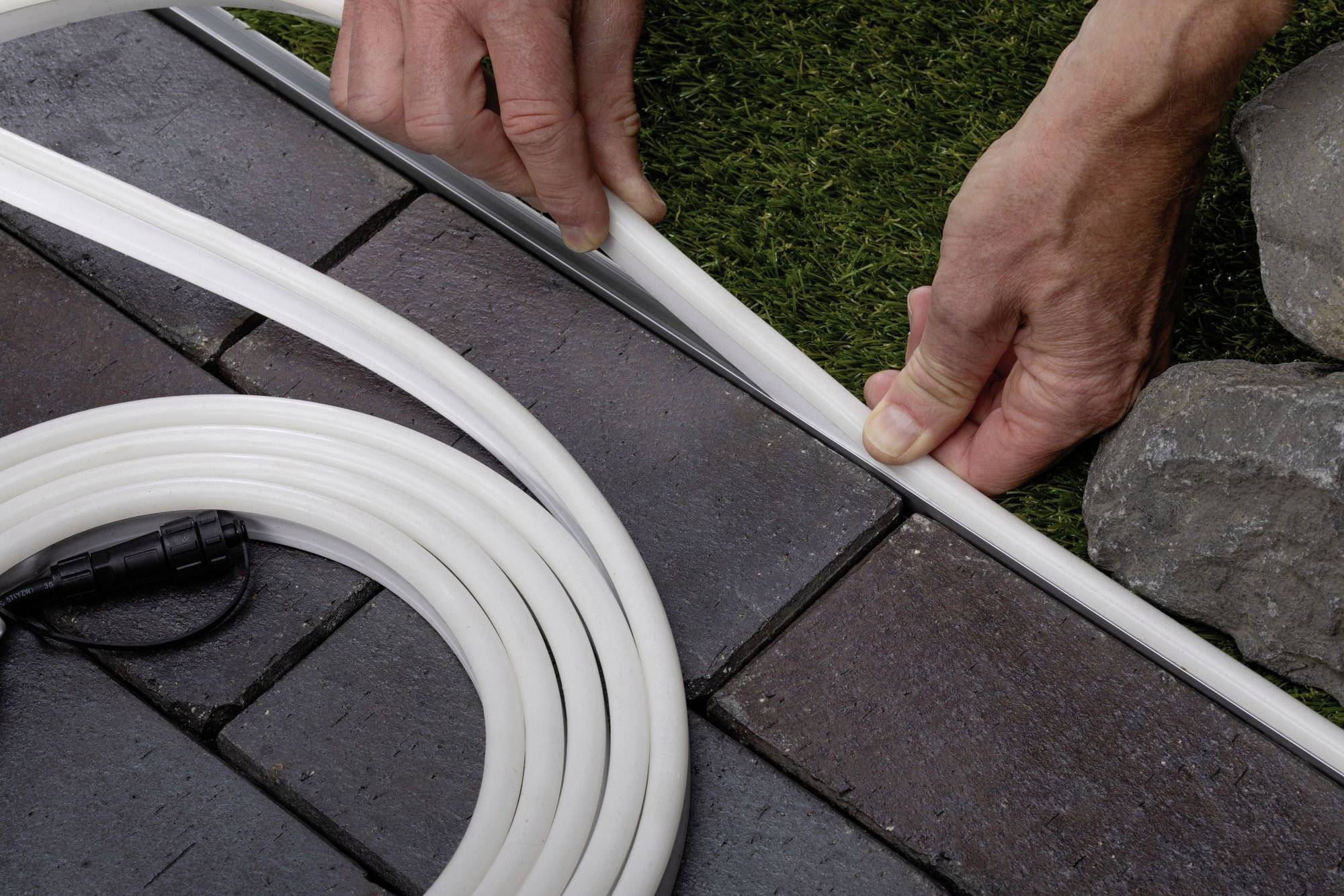 Deux mains installent un tuyau flexible blanc le long d'une surface pavée à côté de l'herbe et des pierres.