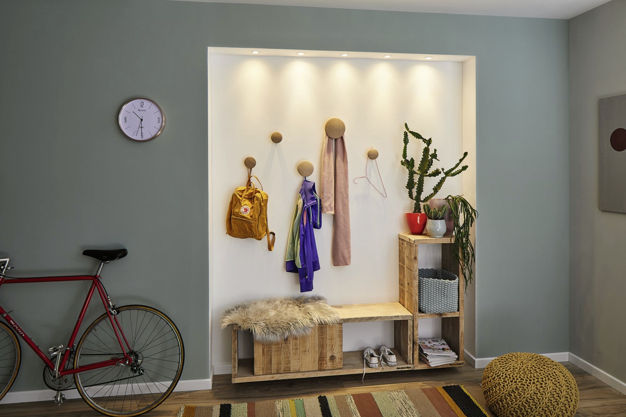 Entrée avec vestiaire en bois : vestes, sac à dos et écharpe suspendus ; plantes décoratives ; vélo accroché au mur ; horloge indiquant 8 heures.