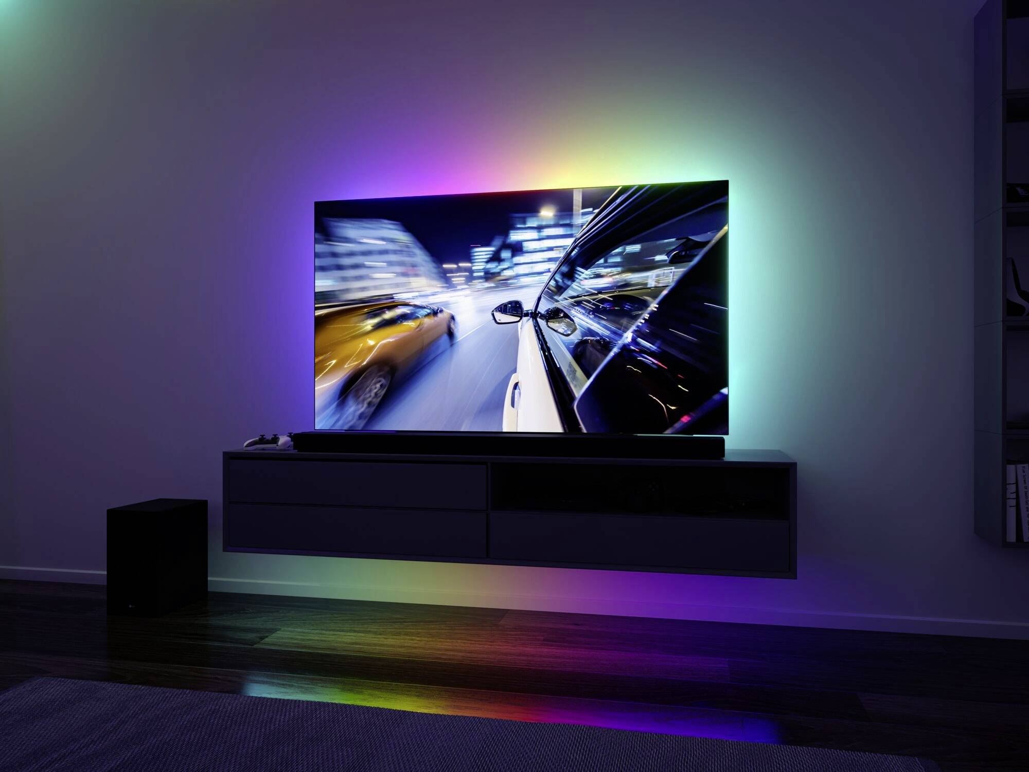 Un salon moderne avec un grand téléviseur au mur. L'écran affiche une scène urbaine floue de nuit. Un éclairage d'ambiance coloré brille derrière la télévision.