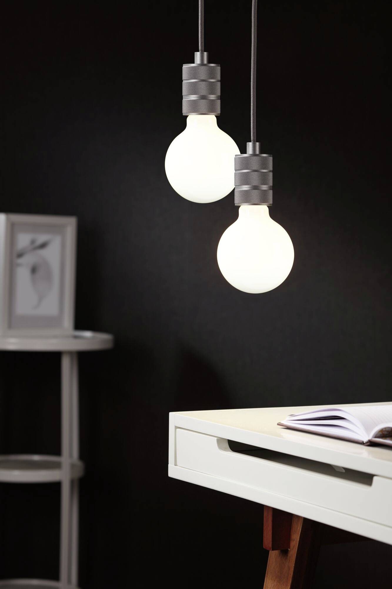 Deux lampes suspendues éclairent un bureau blanc sur lequel un carnet ouvert est posé. Un étagère se trouve en arrière-plan.