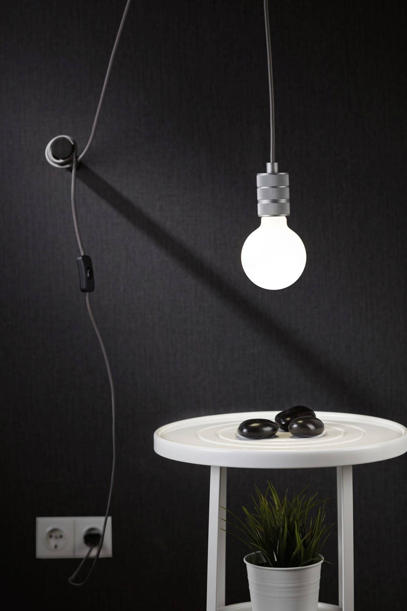 Une ampoule suspendue brille au-dessus d'une table blanche avec des pierres noires et une plante dans un pot.