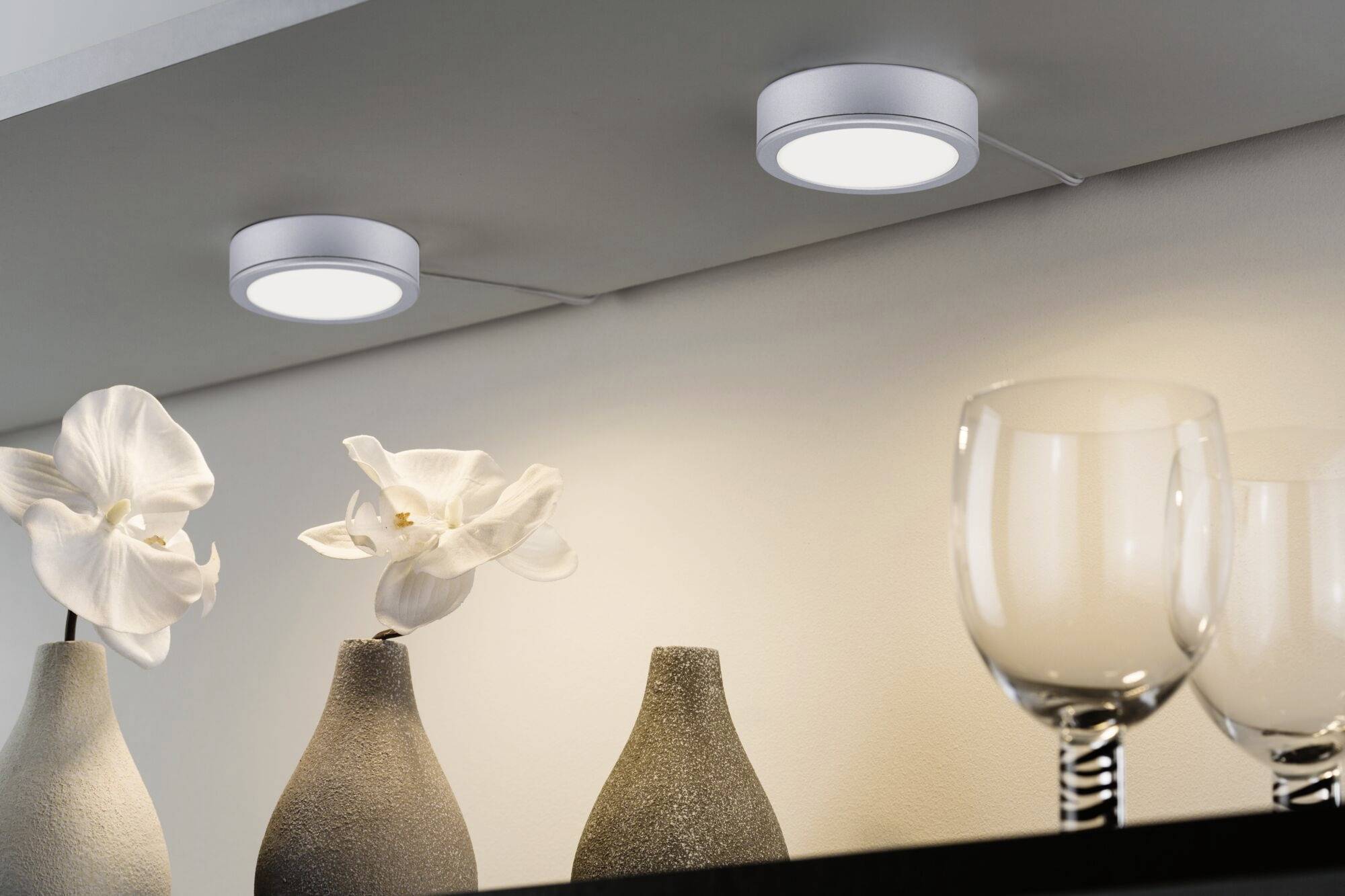 Deux spots de plafond blancs illuminent une scène décorative composée de deux fleurs blanches dans des vases gris et de deux verres à vin transparents posés sur une étagère.
