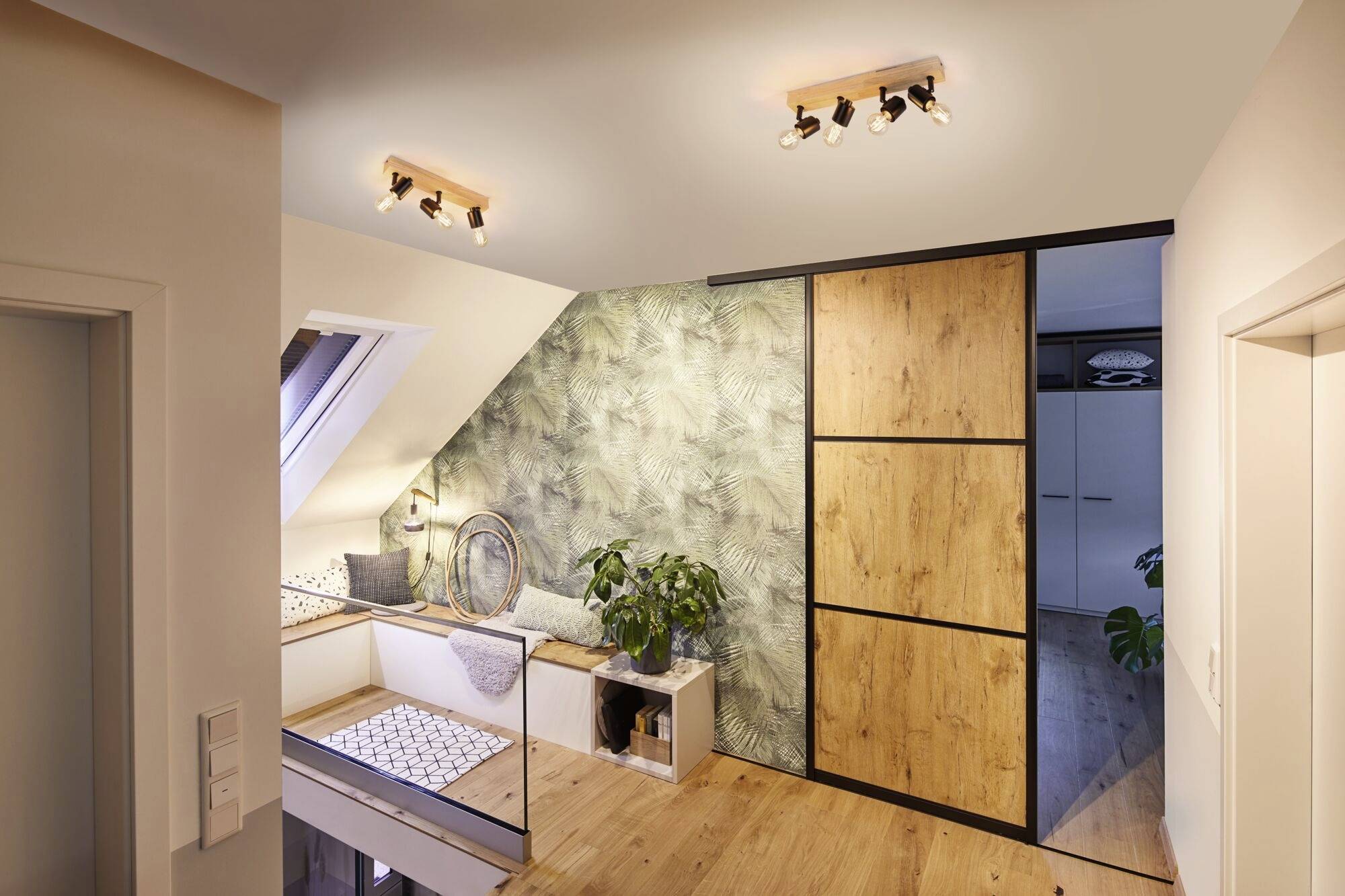 Combles avec un sol en bois clair, banc sous une fenêtre de toit, plante et armoire à portes en bois. Papier peint à motifs sur le mur.