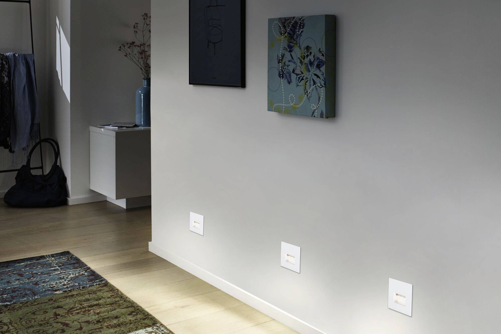 Un couloir lumineux avec un sol en bois, un tapis et deux œuvres d'art accrochées au mur. À gauche, se trouve une petite table avec des vases.