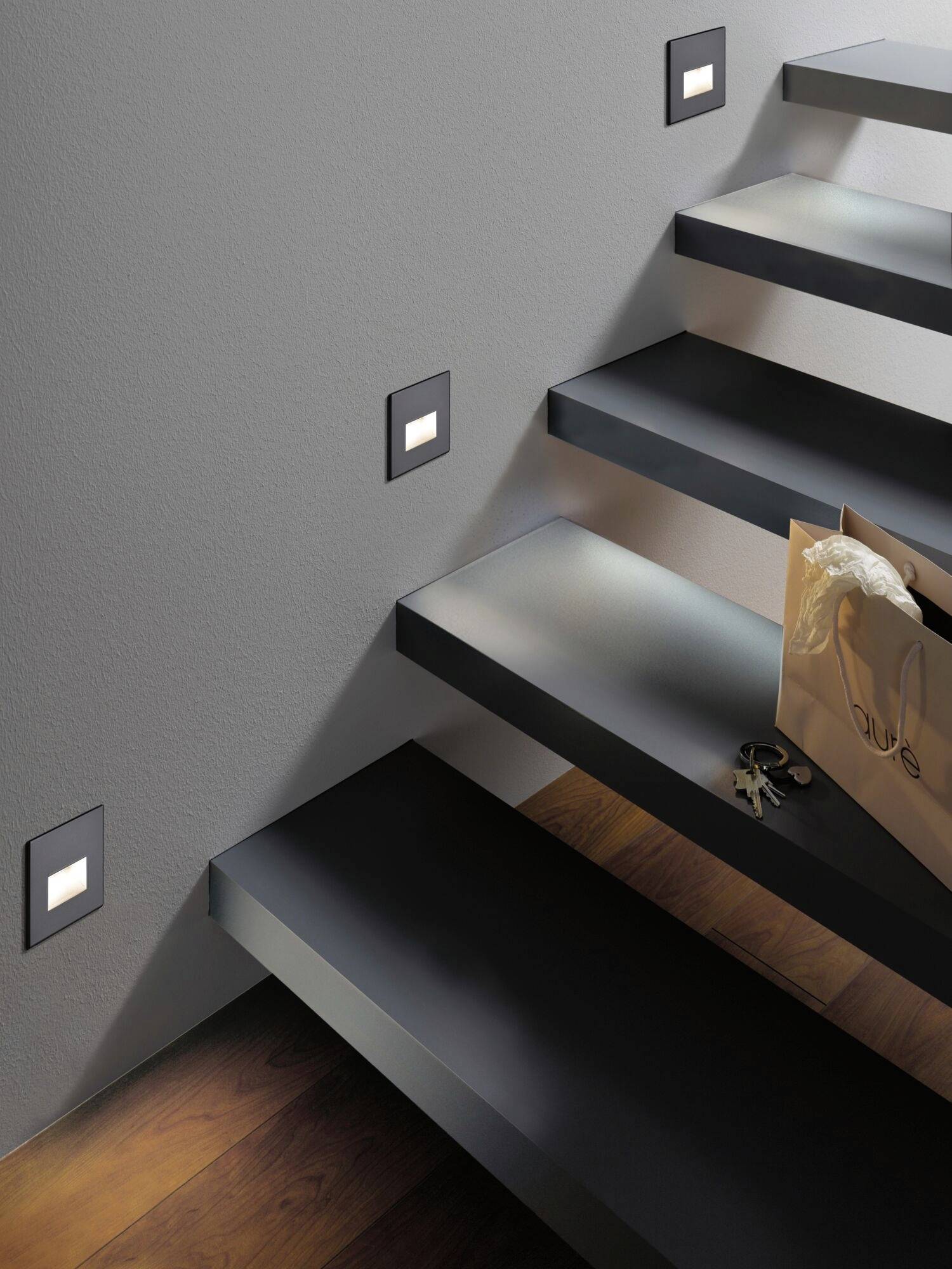 Escalier avec un éclairage moderne ; à droite, un sac contenant des clés et des lunettes posé sur une marche. Les marches sombres contrastent avec le mur clair.