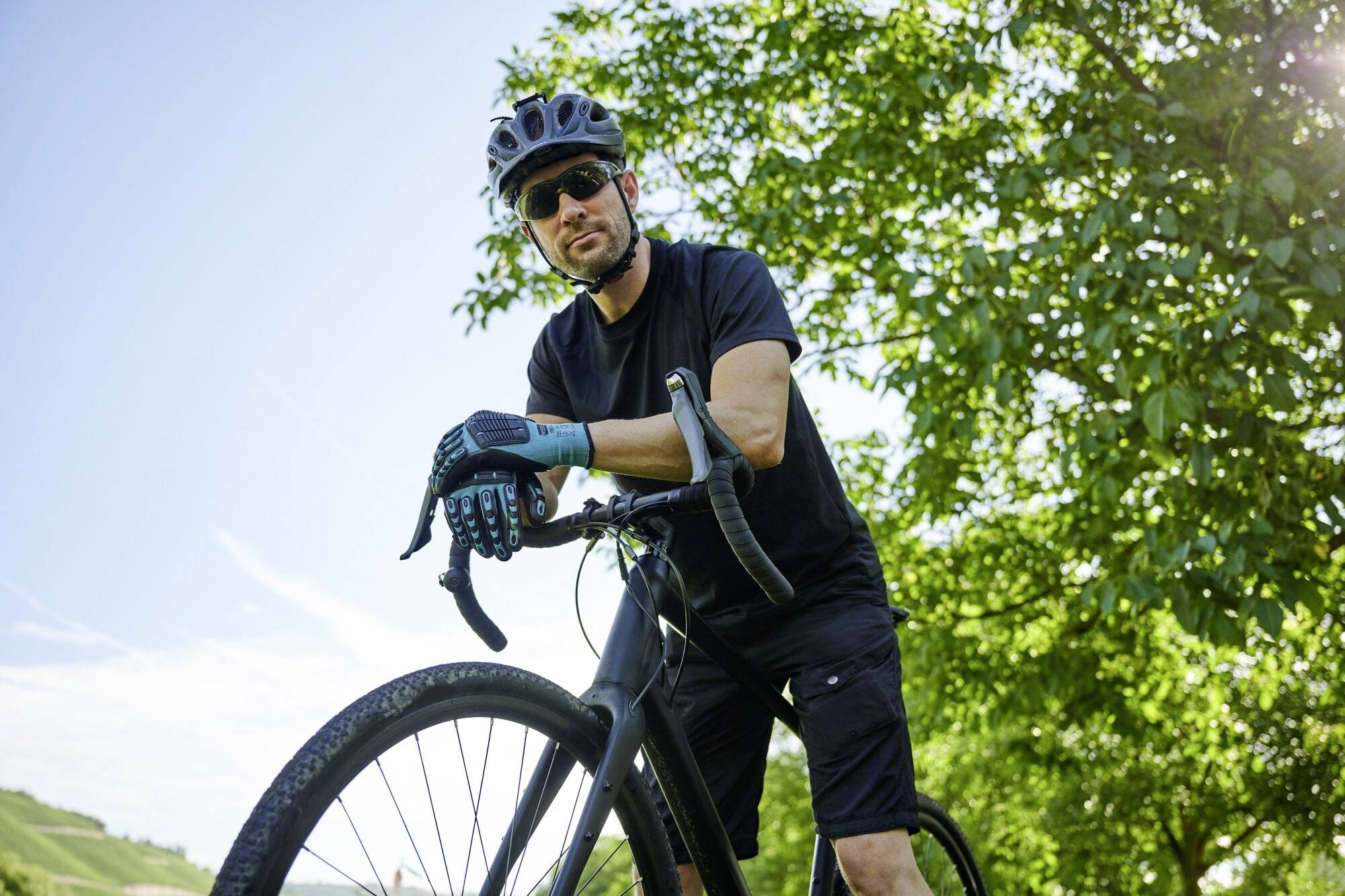 Un homme en tenue de sport pose avec un vélo de course dans un environnement verdoyant, par temps ensoleillé. Il porte un casque et des lunettes de soleil.