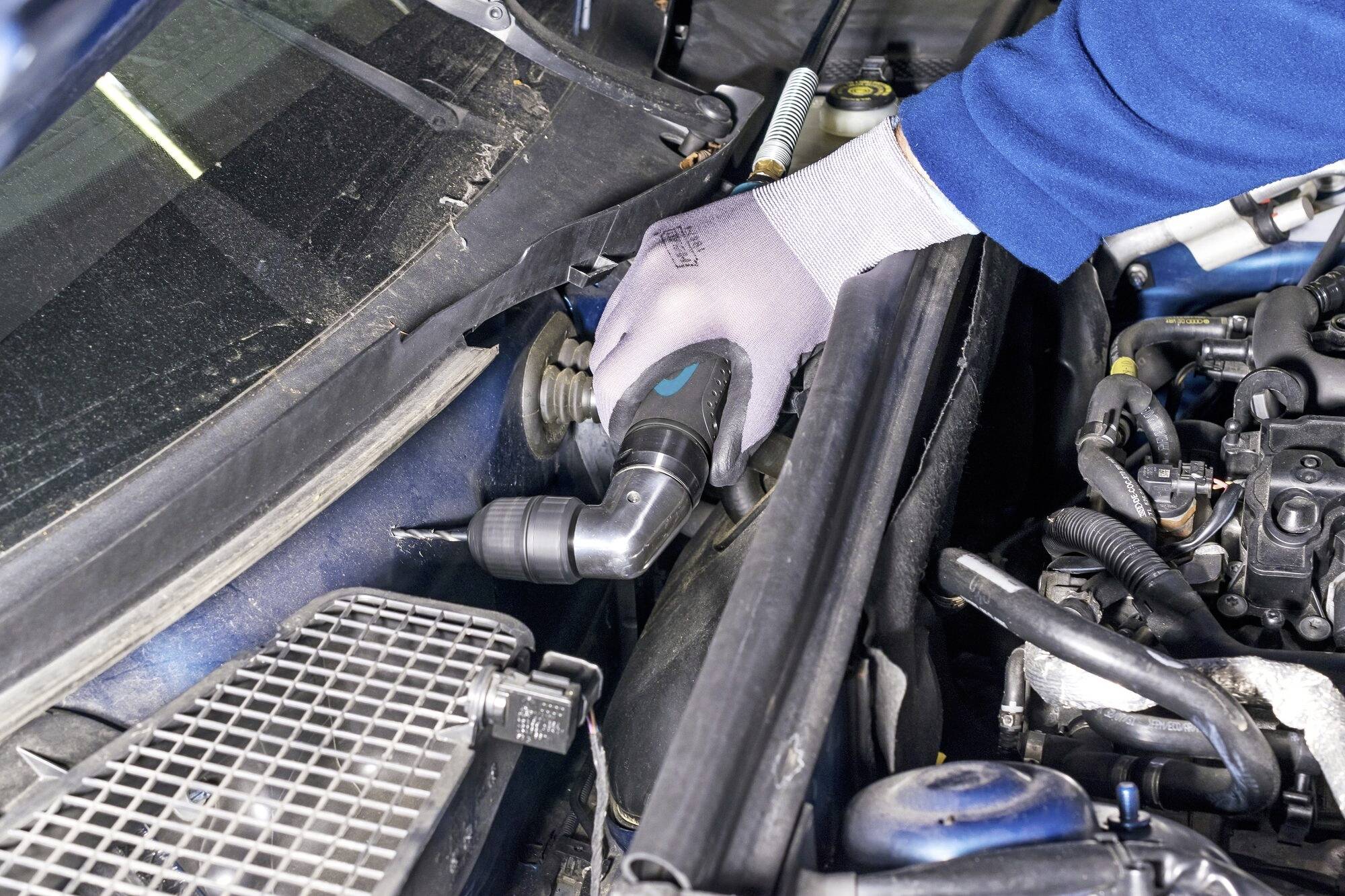 Un mécanicien portant des gants bleus utilise un outil pour réparer le moteur d'un véhicule. Le compartiment moteur est partiellement visible.