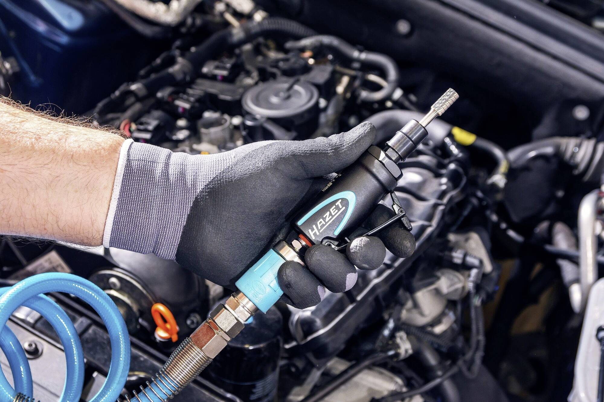 Une main gantée tient un outil de ponçage pneumatique au-dessus d'un moteur automobile partiellement assemblé. Un tuyau bleu est connecté.