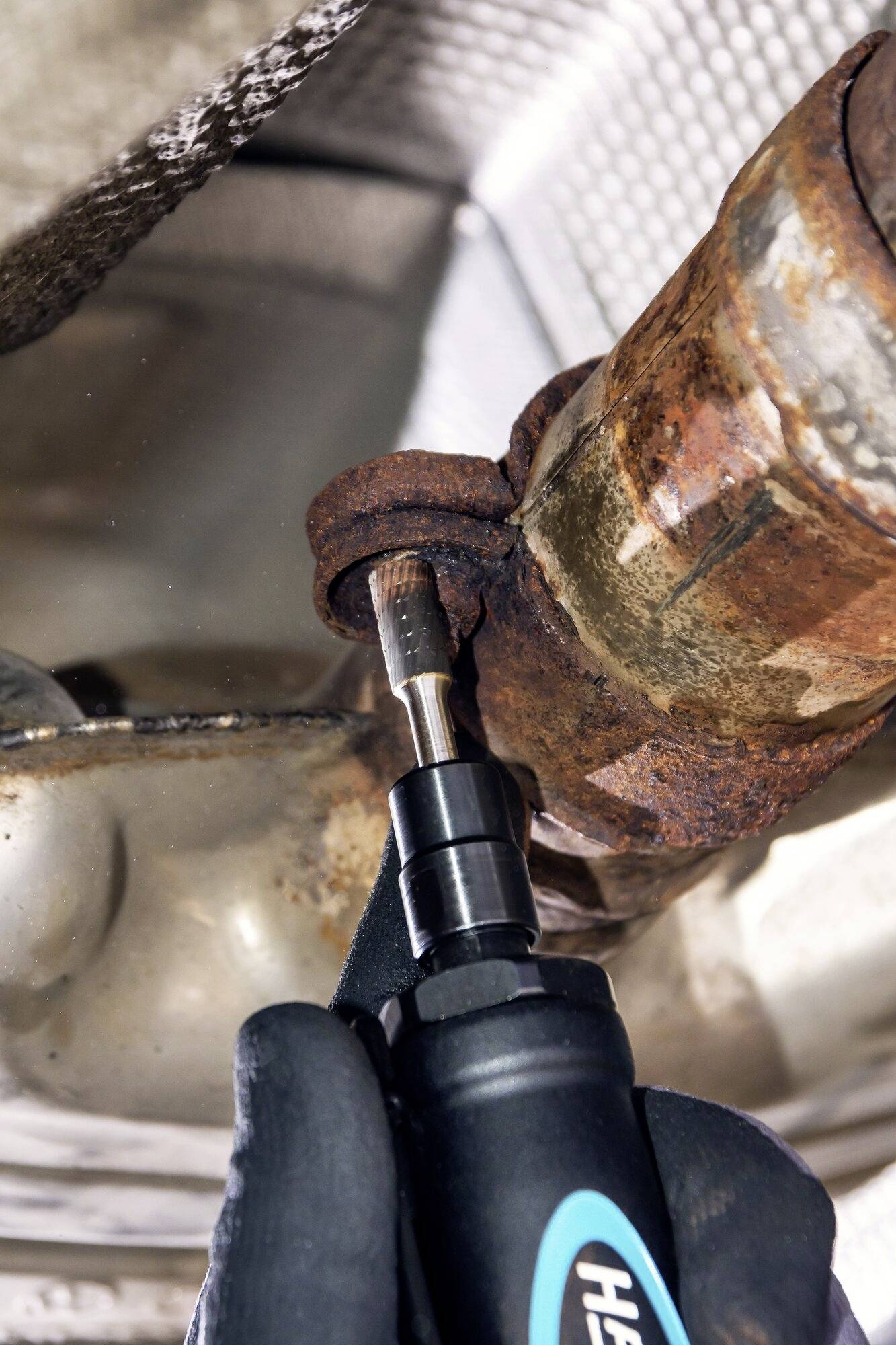 Une main tient un outil pour desserrer une vis rouillée sur une charnière métallique. Situation de maintenance industrielle.