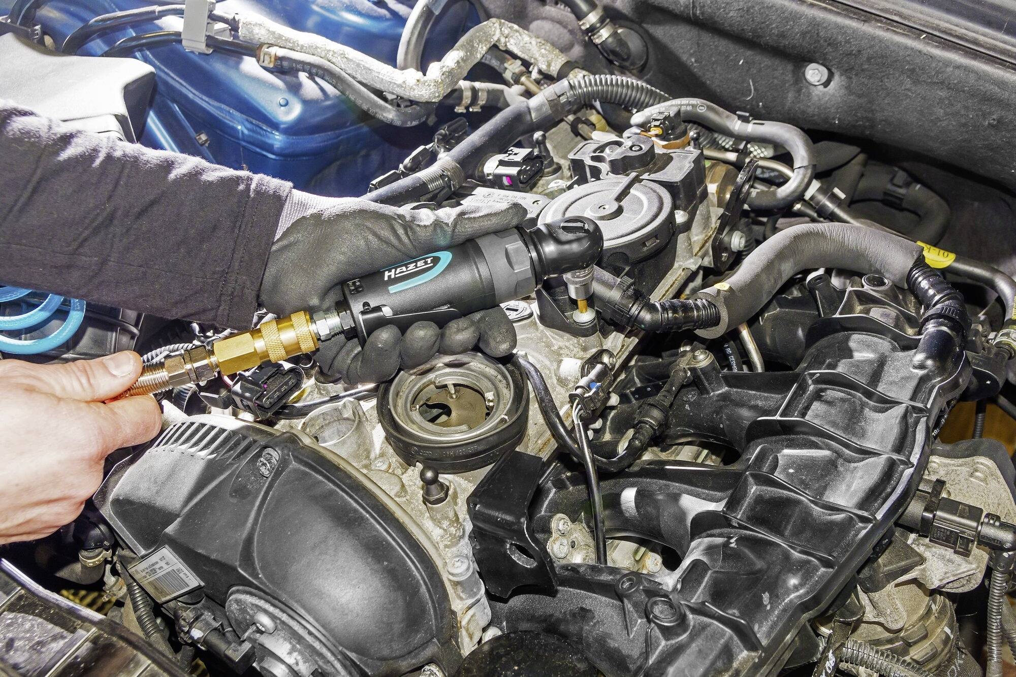 Un mécanicien répare un moteur de voiture à l'aide d'un outil spécial, entouré de différents composants et tuyaux du moteur.