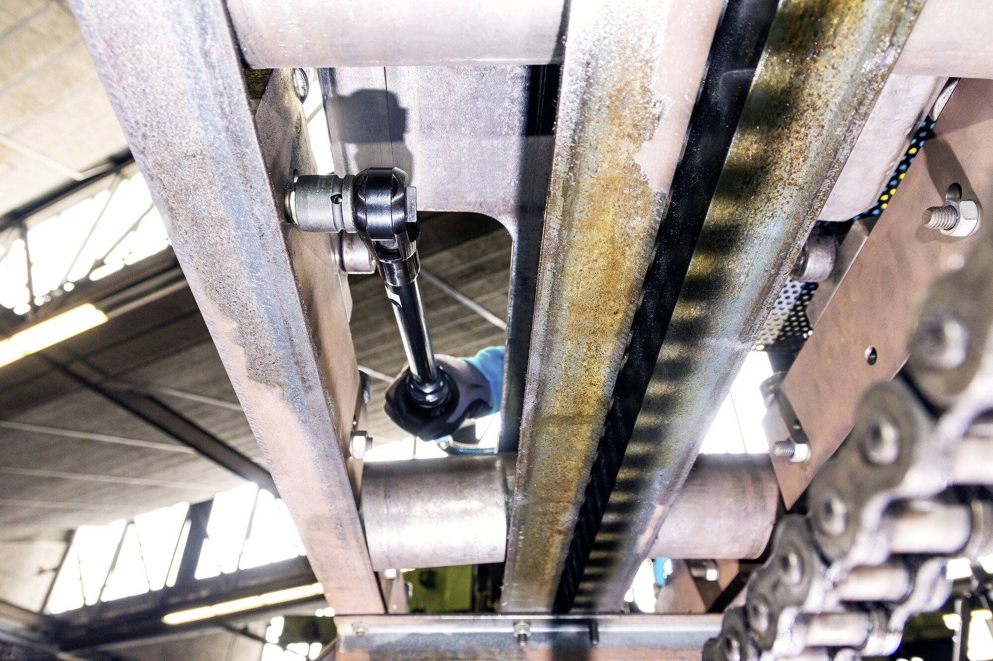 Vue d'une machine industrielle depuis le dessous, un ouvrier la répare à l'aide d'un outil. Chaînes et structures métalliques visibles.