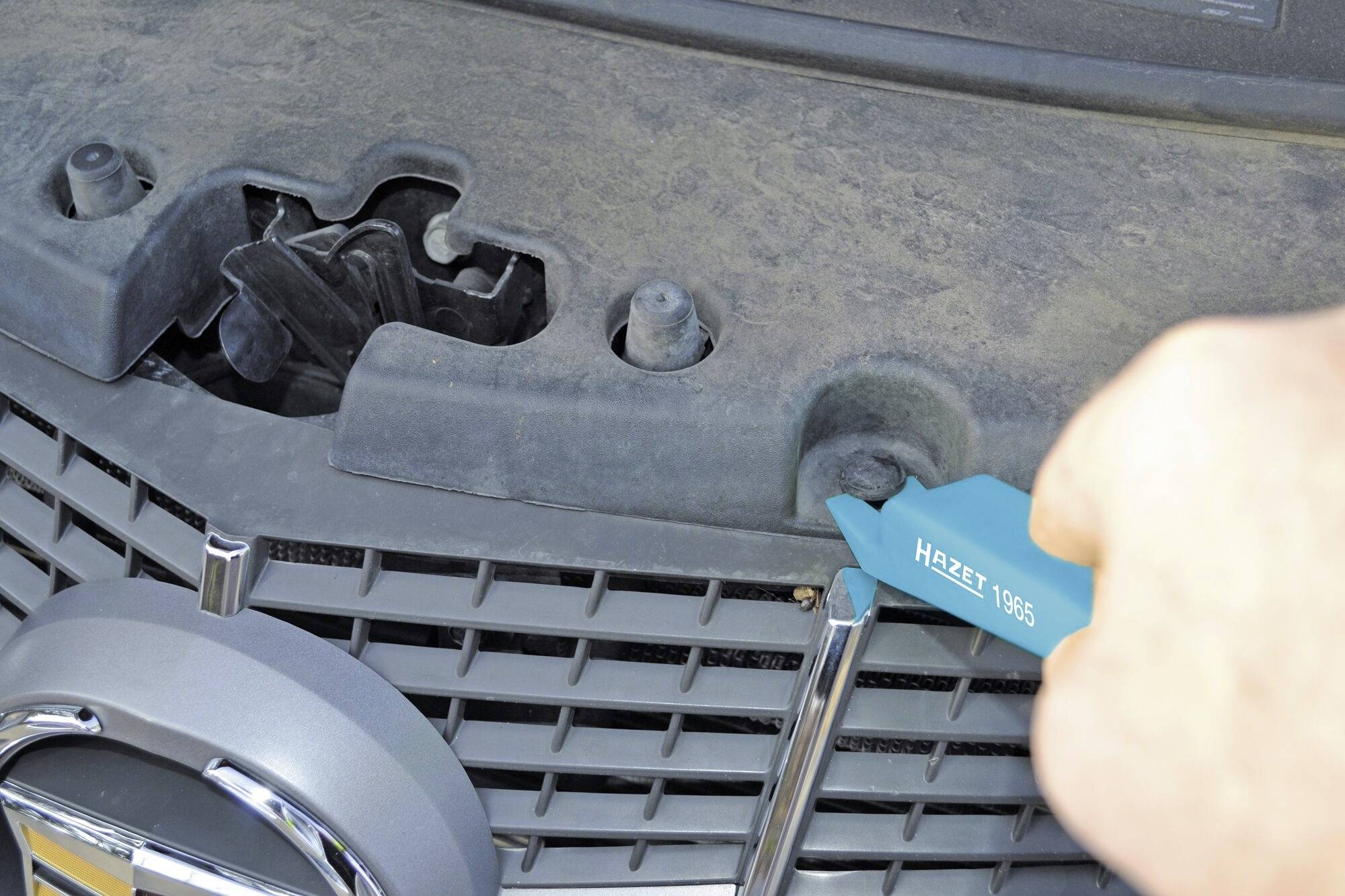 Une main manipule un outil en plastique bleu pour détacher un clip sur une grille de voiture sale.