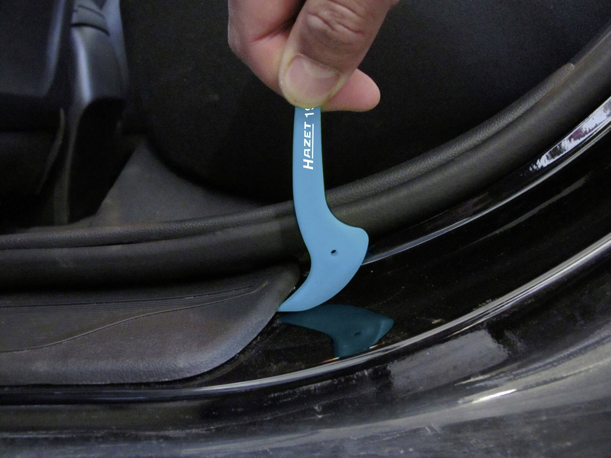 Une main utilise un outil en plastique bleu pour retirer un habillage de pièce automobile. L'outil porte l'inscription 'Hazet'.