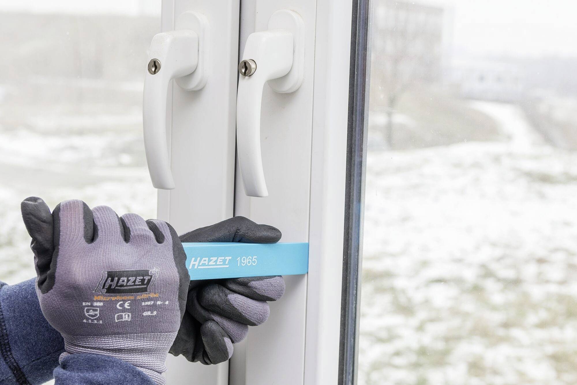 Une personne portant des gants ouvre une fenêtre avec un outil bleu sur un fond enneigé.
