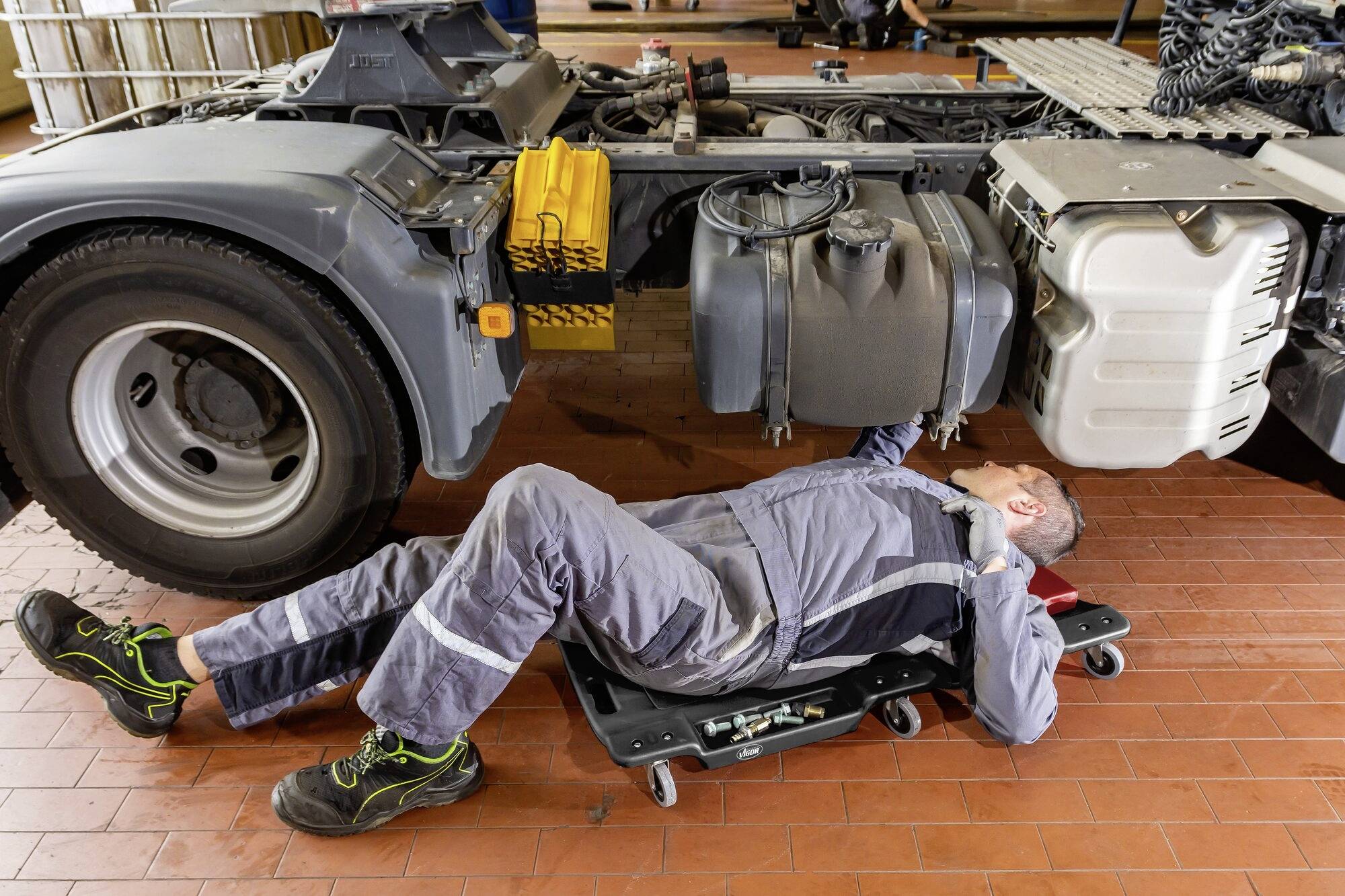 Un mécanicien est allongé sur un chariot de visite sous un camion et effectue des travaux de maintenance. Il porte une combinaison grise et des chaussures de travail.