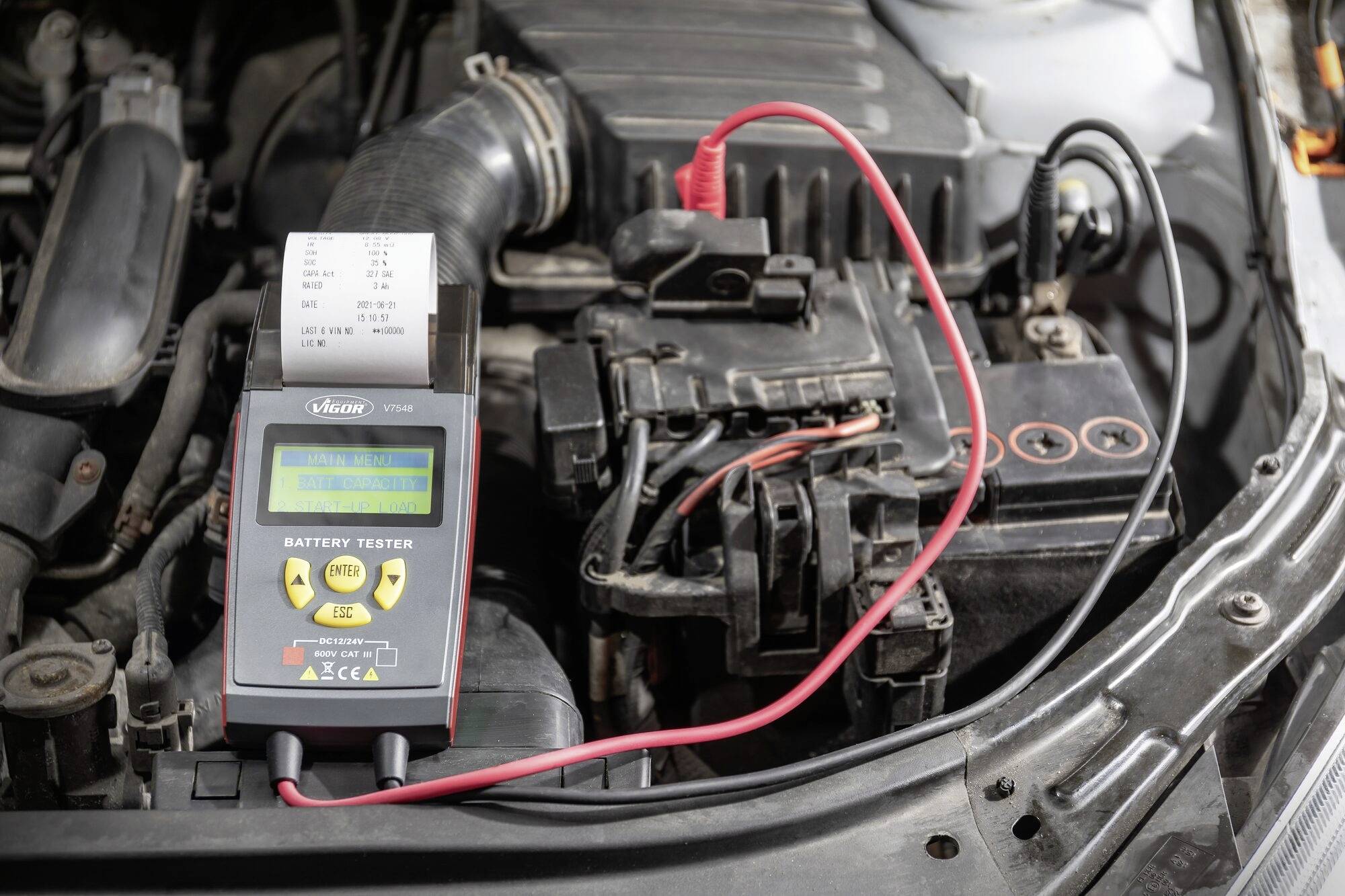 Testeur de batterie sur le capot ouvert d'une voiture affichant les résultats. Le câble rouge est connecté à la batterie.