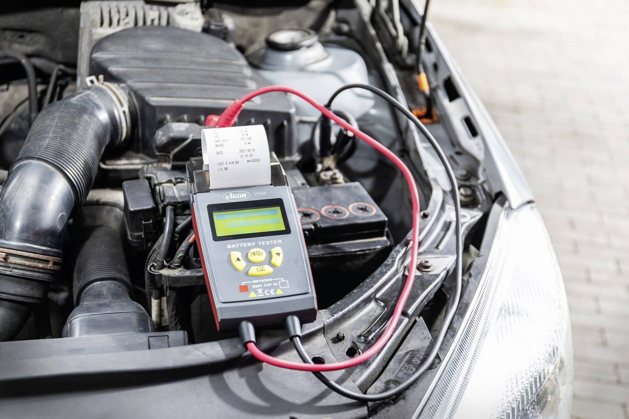 Un testeur de batterie de voiture est connecté à la batterie d'un compartiment moteur ouvert. L'appareil affiche les résultats sur un écran.