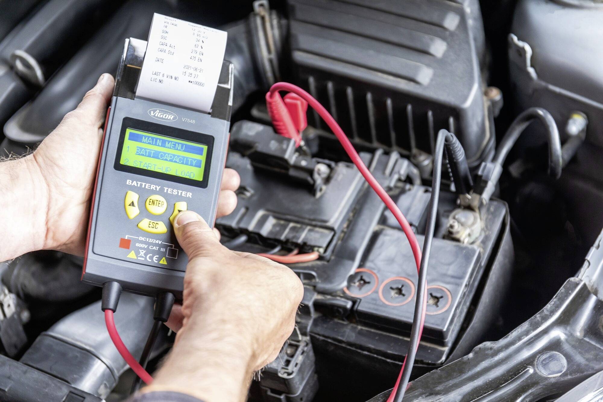 Une personne teste une batterie de voiture à l'aide d'un testeur de batterie numérique. Un câble est connecté à la batterie et l'écran affiche les résultats.