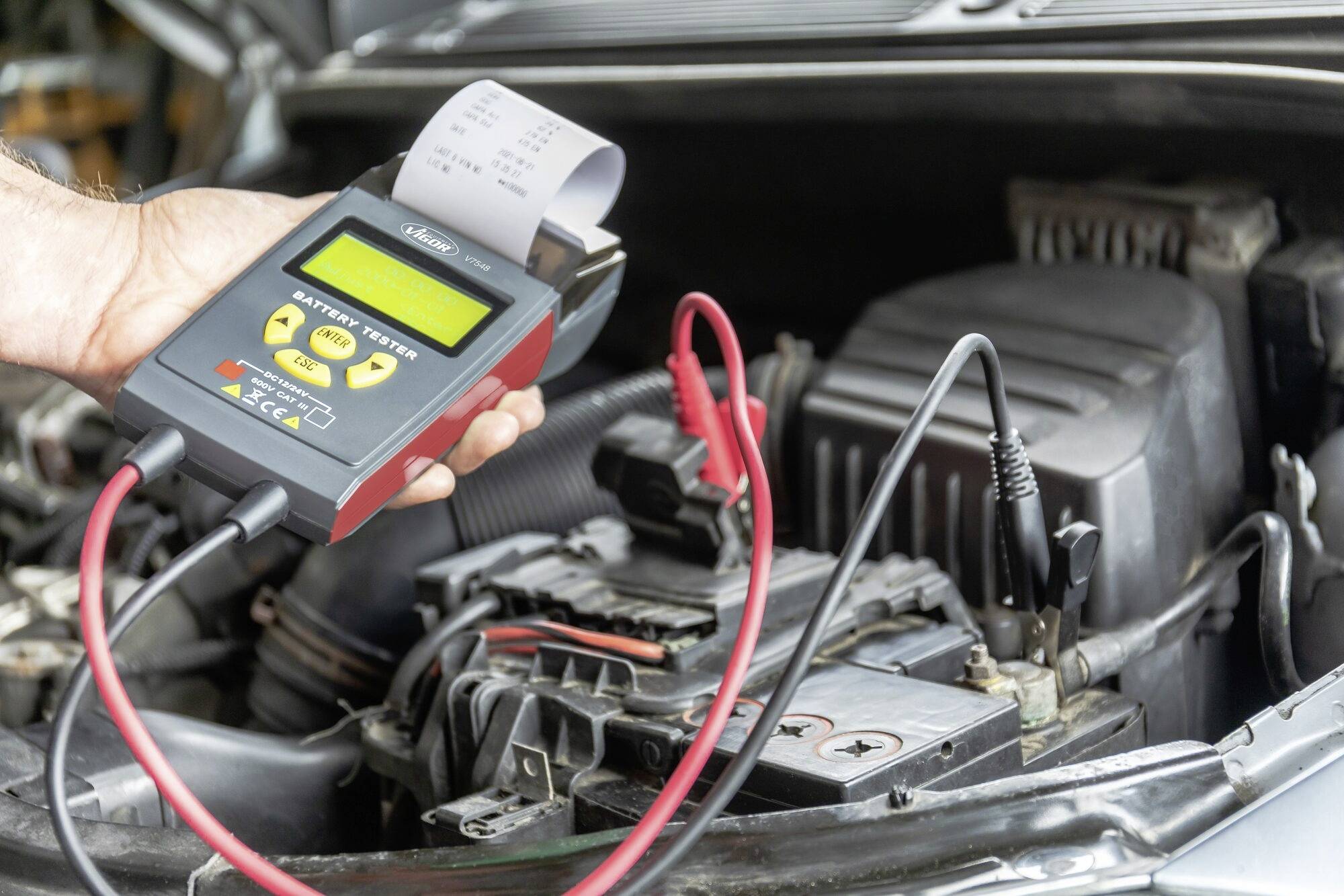 Une main tient un dispositif de test de batterie connecté à une batterie de voiture. L'écran affiche des valeurs de mesure, et une bande de papier est imprimée.