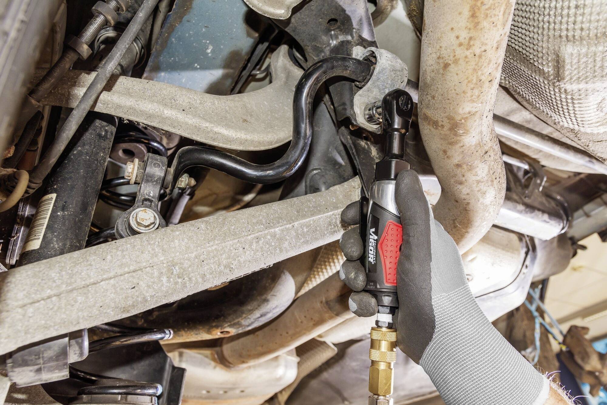 Main d'un mécanicien serrant une vis avec un visseuse à choc électrique dans un châssis automobile. Vue sur le soubassement et les pièces du véhicule.