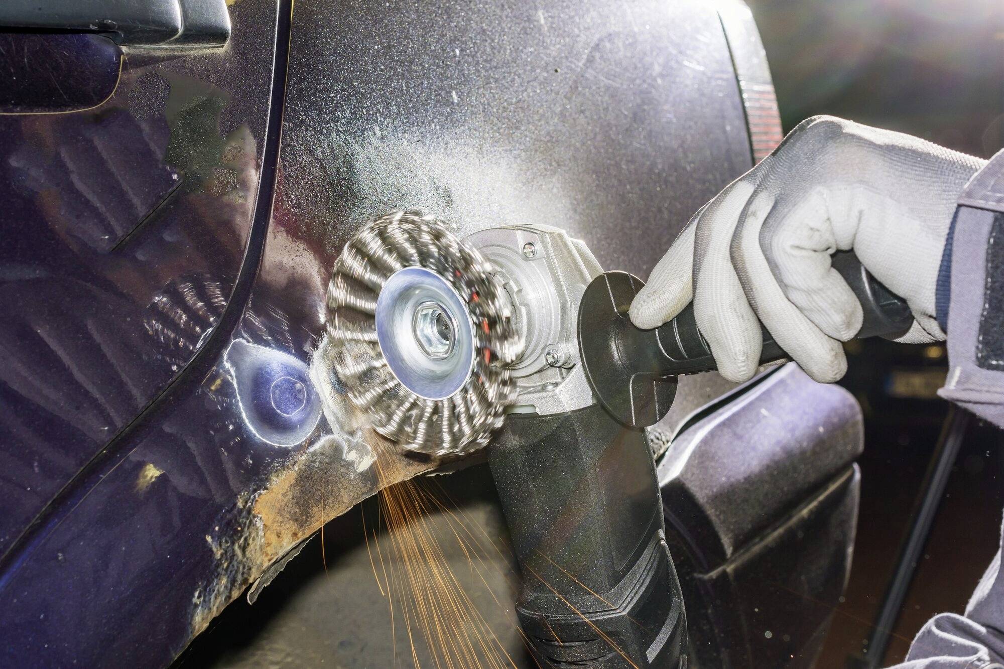 Quelqu'un ponce l'aile rouillée d'une voiture avec une meuleuse d'angle, des étincelles volent. Des mains portent des gants de protection.