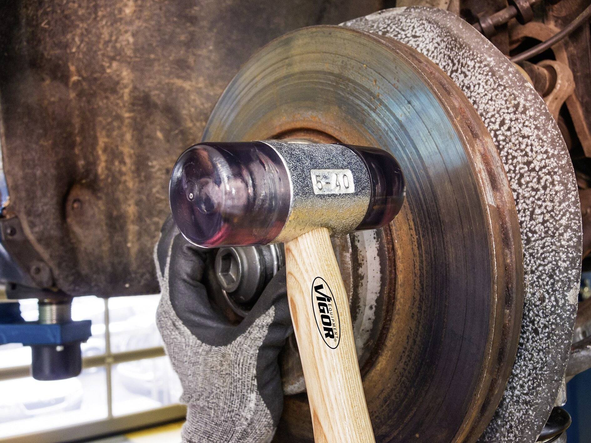Une personne portant des gants utilise un marteau sur un disque de frein rouillé. L'accent est mis sur l'opération de réparation ou de maintenance.