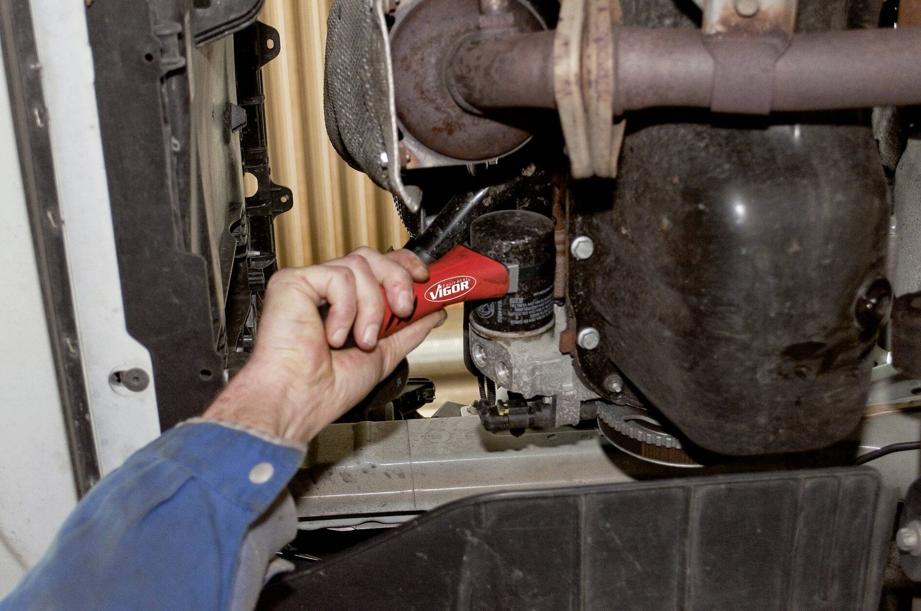 Une personne fait tourner un filtre à huile dans le moteur d'un véhicule à l'aide d'un outil rouge.
