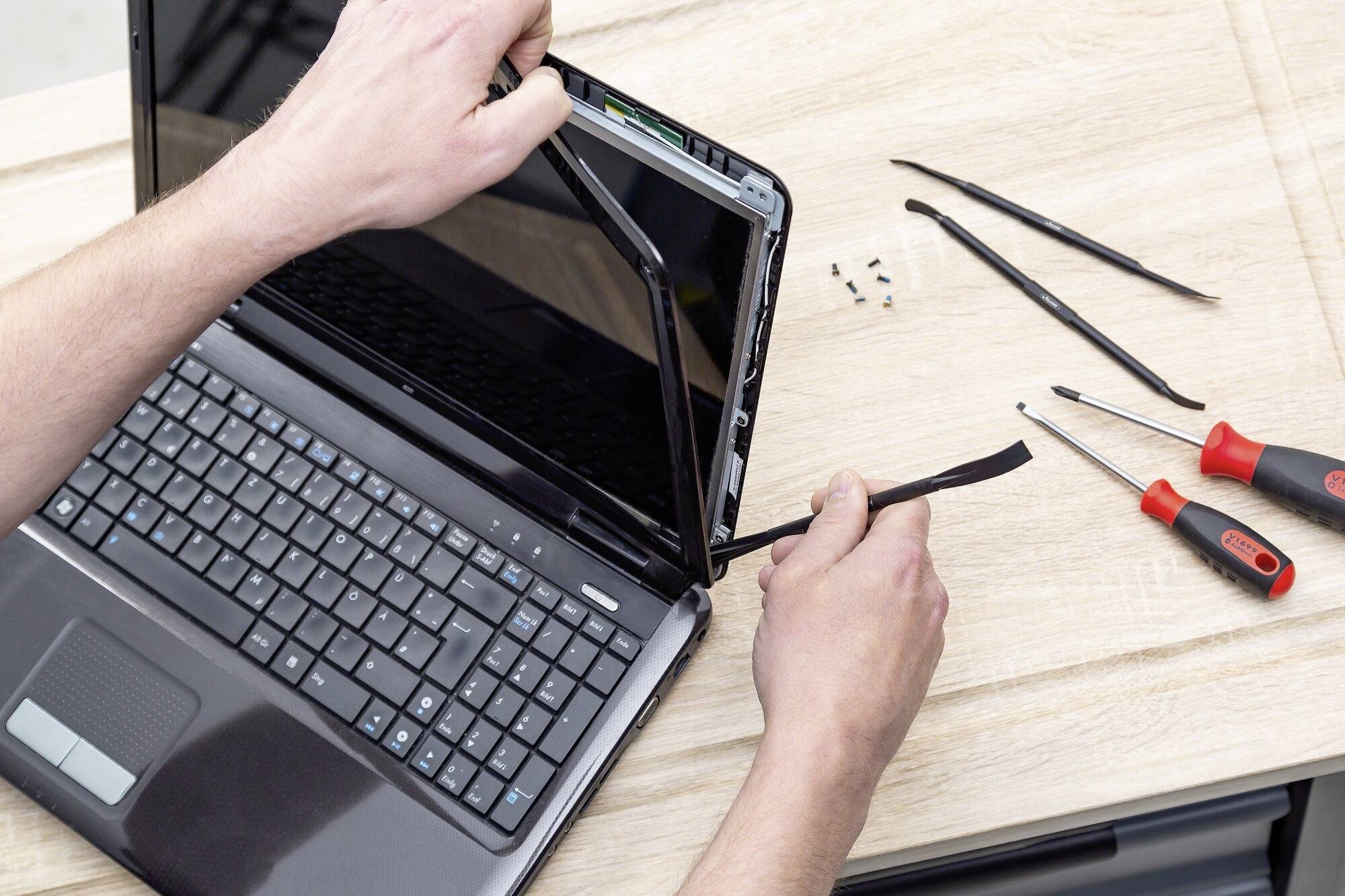Une personne répare un ordinateur portable en retirant le cadre de l'écran. Des outils et des vis sont disposés sur la table.