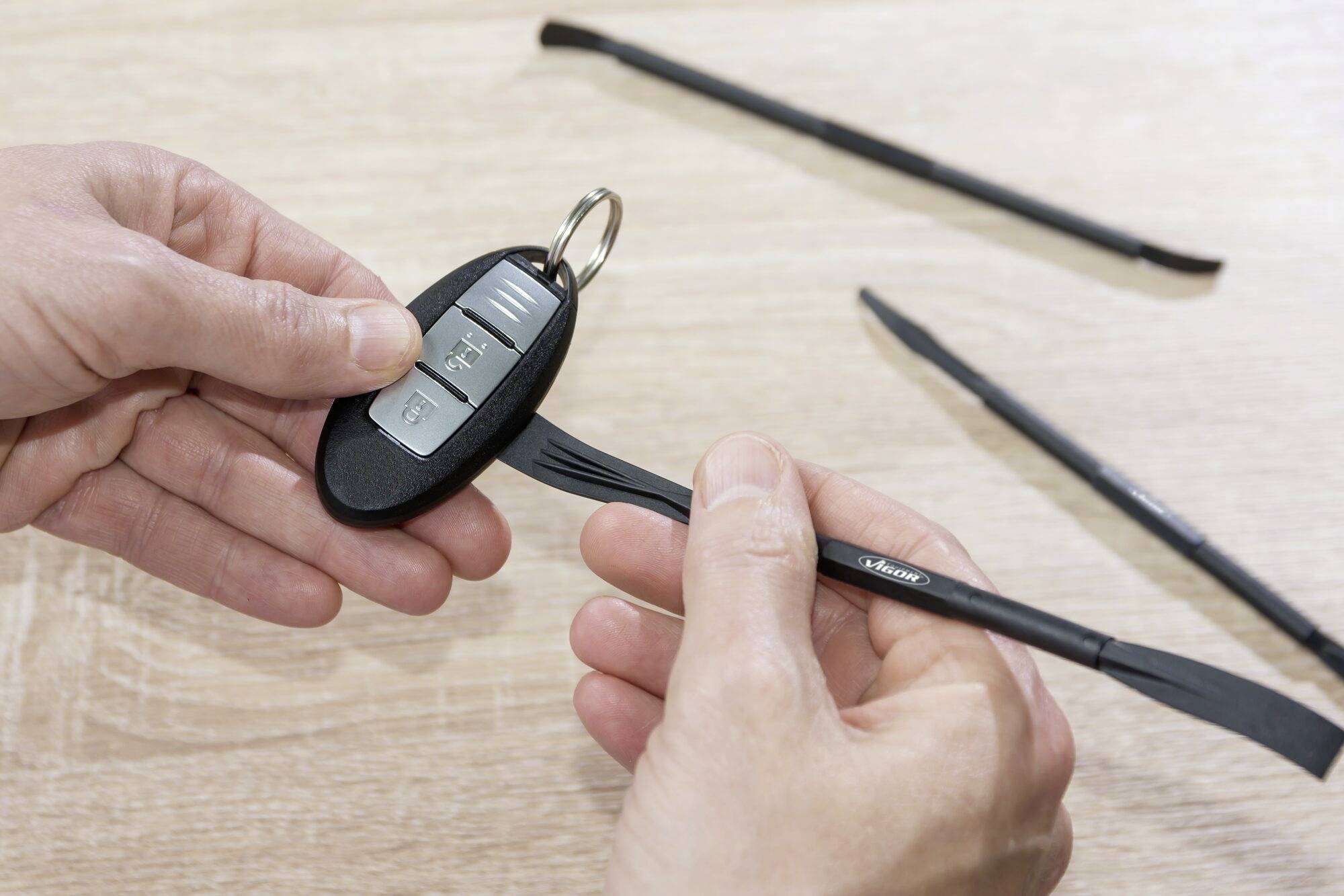 Gros plan sur deux mains tenant un outil en plastique et une clé de voiture avec des boutons. D'autres outils sont posés sur une table en bois.