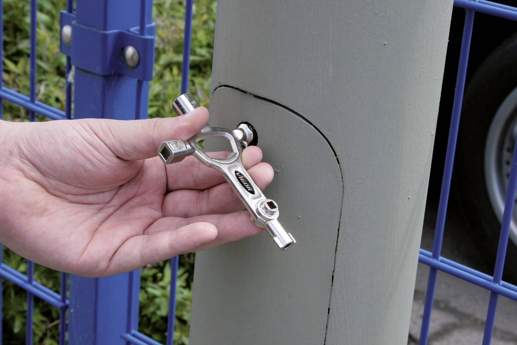 Une main tient une clé de vanne qui est utilisée sur le mécanisme de verrouillage d'un pilier gris, près d'une clôture métallique bleue.