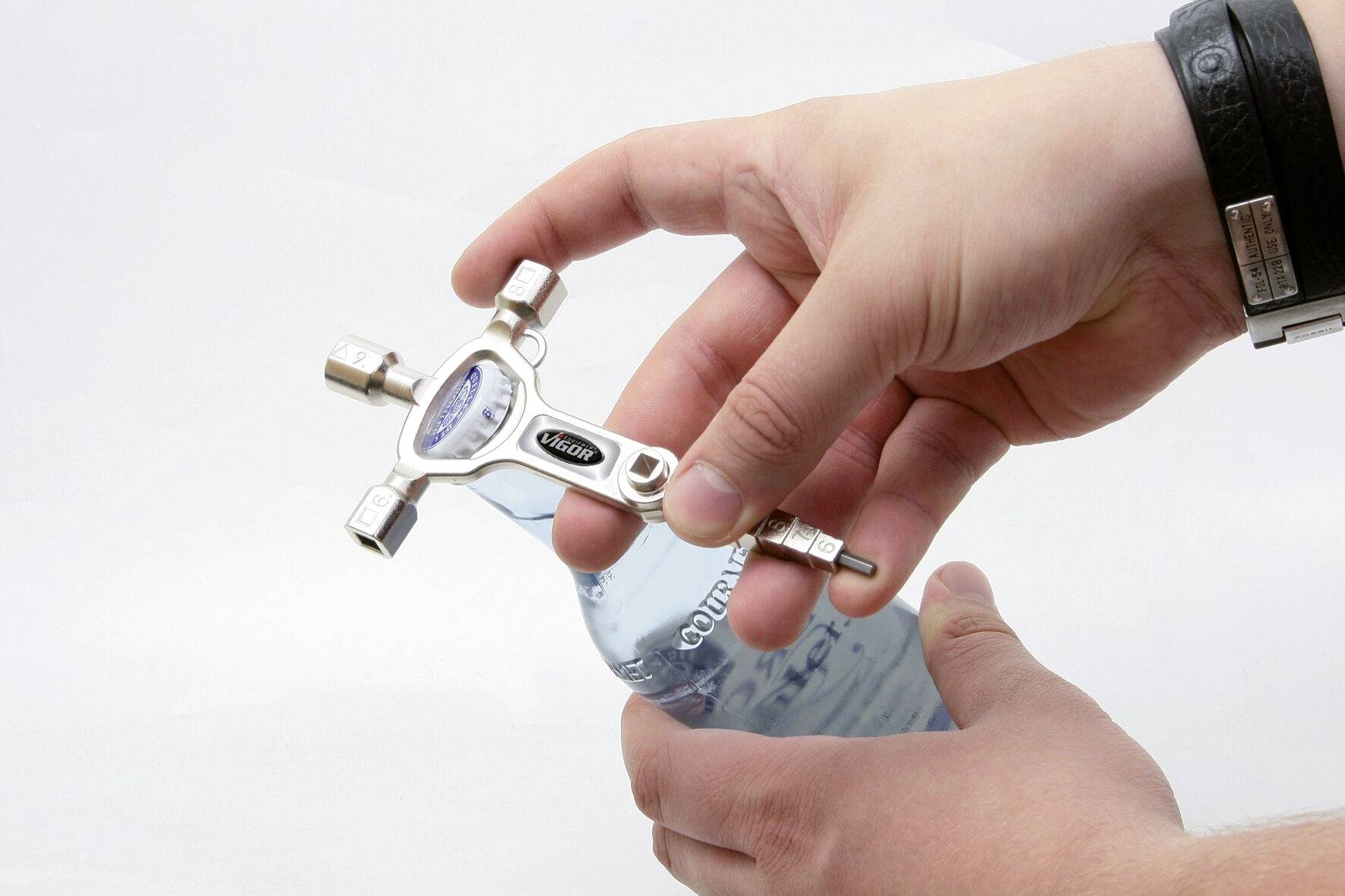Une personne tient un outil multifonction avec plusieurs douilles et l'utilise pour ouvrir une bouteille en verre partiellement visible.