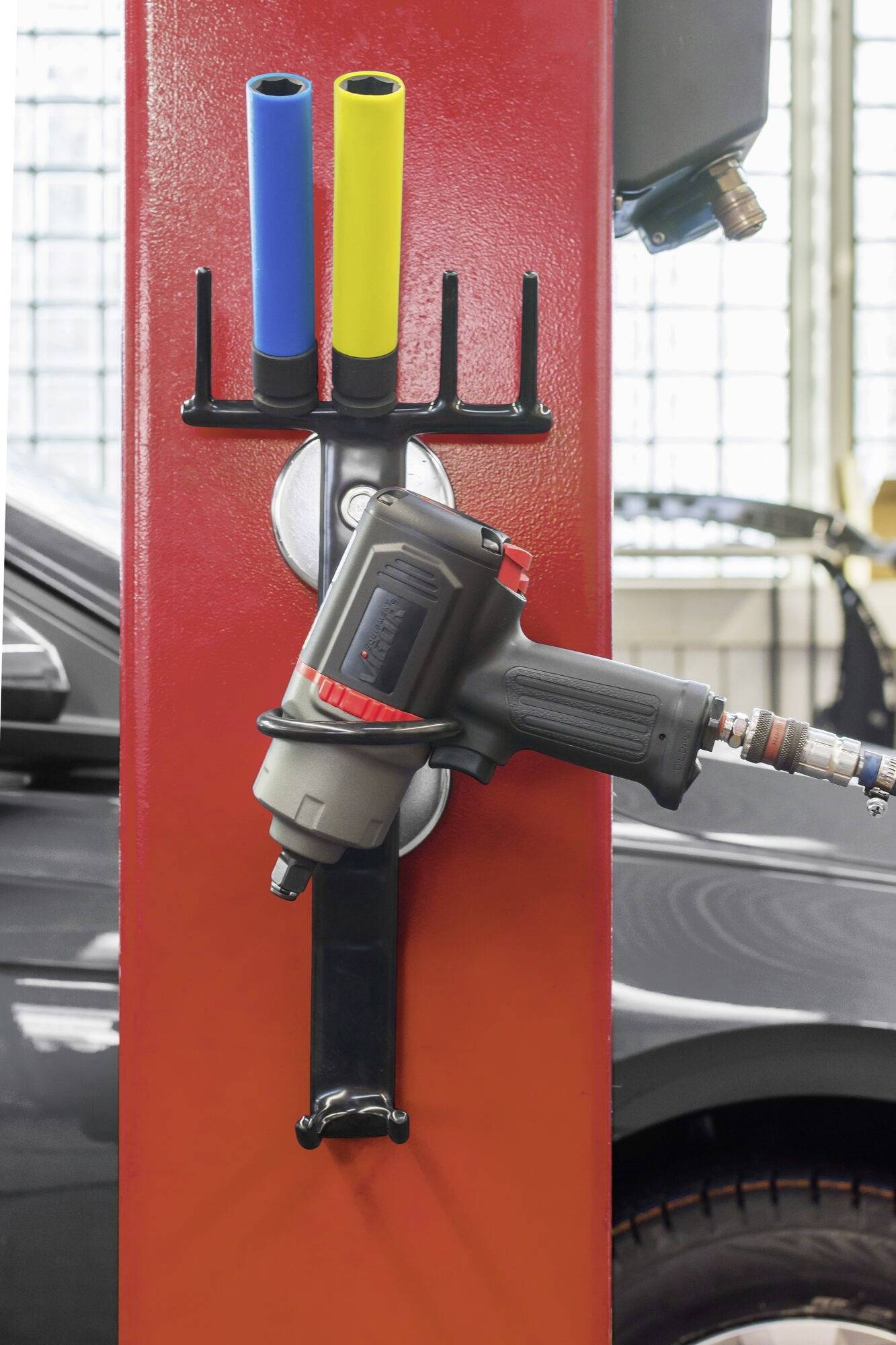 'Un tournevis à choc est suspendu à un support rouge dans un atelier automobile, entouré de trois tuyaux pneumatiques colorés ; une voiture est visible en arrière-plan.'