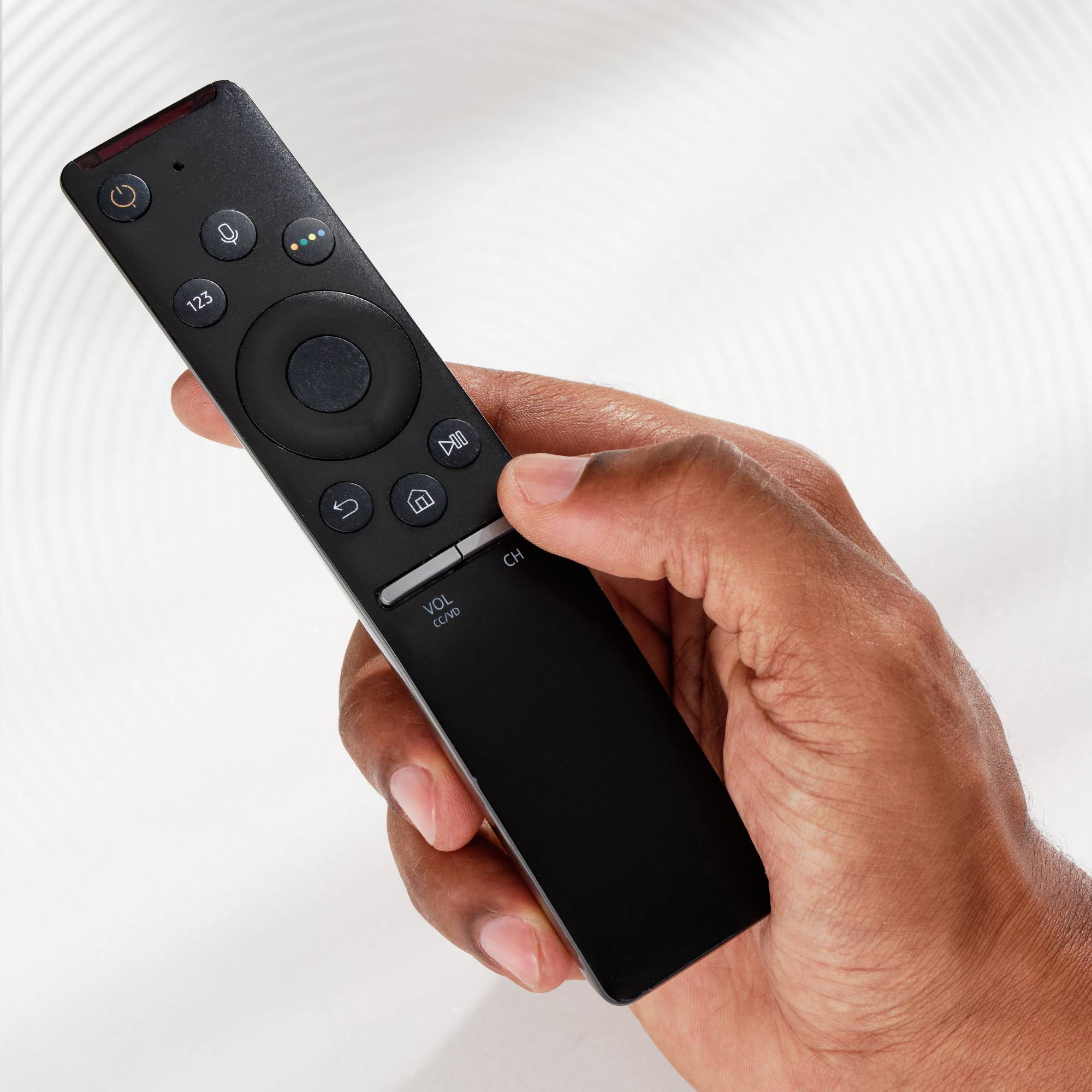 Une main tient une télécommande noire avec plusieurs touches, dont un régulateur de volume et un bouton de changement de chaîne.