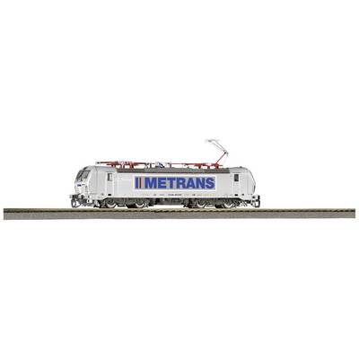 Une locomotive grise portant l'inscription 'METRANS' roule sur des rails. Elle est moderne et équipée de lignes électriques.