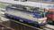 Une locomotive bleue et crème est stationnée sur une voie dans une gare. À l'arrière-plan, on peut voir d'autres trains et quais.