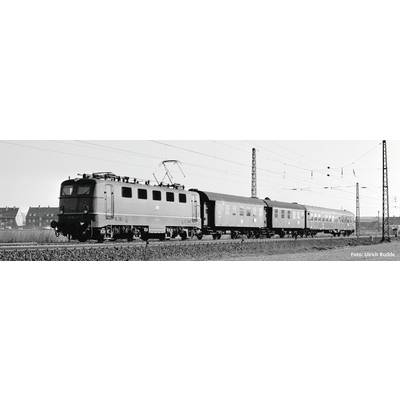 Photo noir et blanc d'un train avec une locomotive et deux wagons sur des rails, des lignes électriques au-dessus, des maisons en arrière-plan.