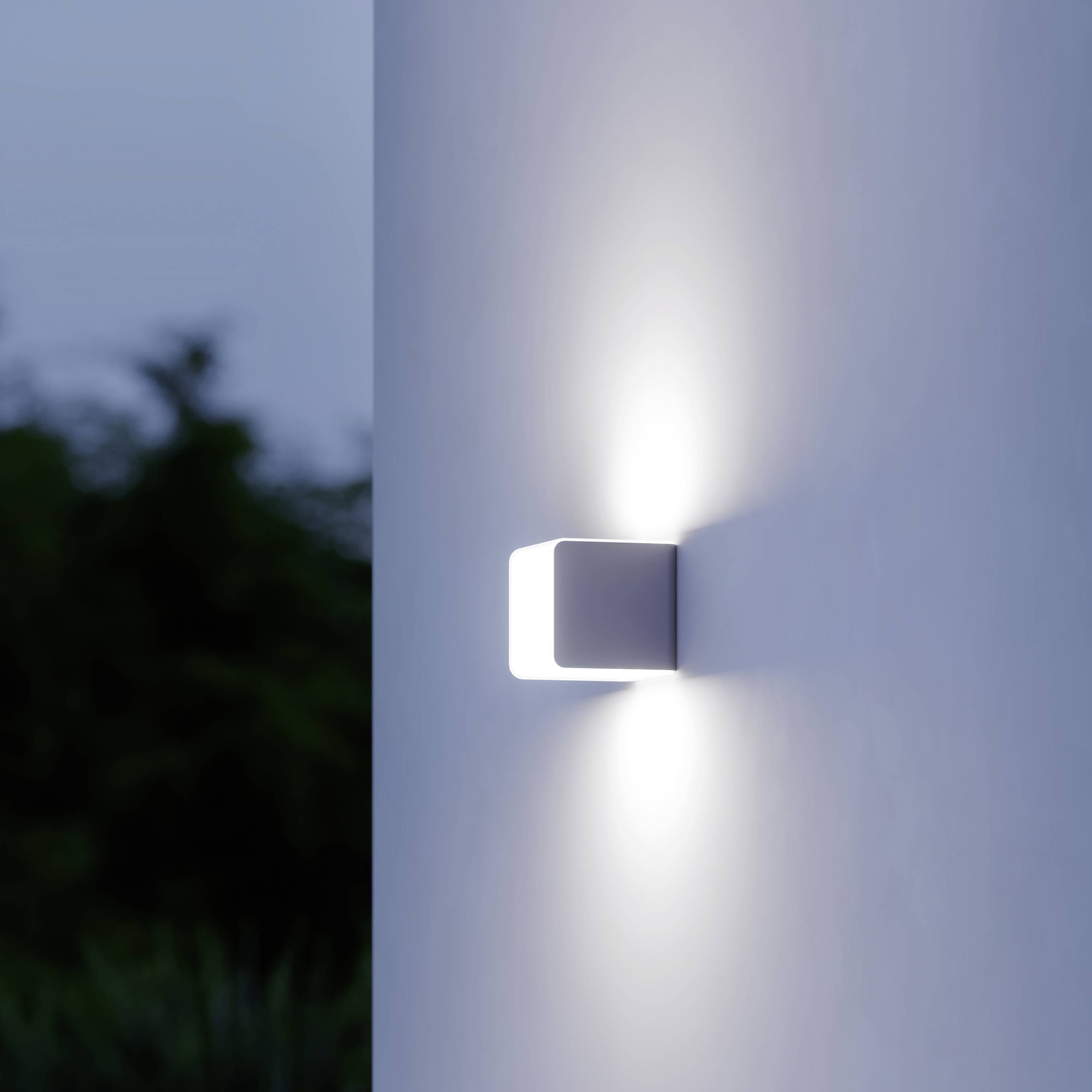 Un luminaire cubique projette une lumière douce sur un mur blanc dans un environnement crépusculaire. Des arbres flous sont visibles en arrière-plan.